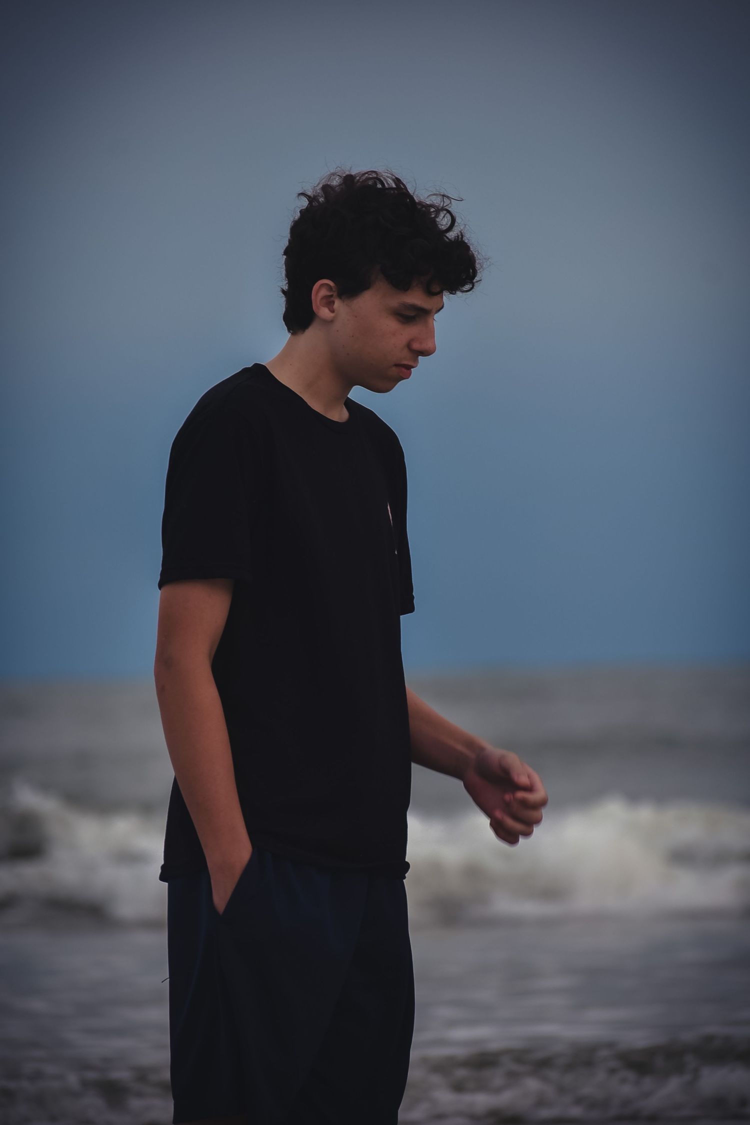 Young man in black t-shirt standing on a beach with ocean waves, looking down pensively at dusk.