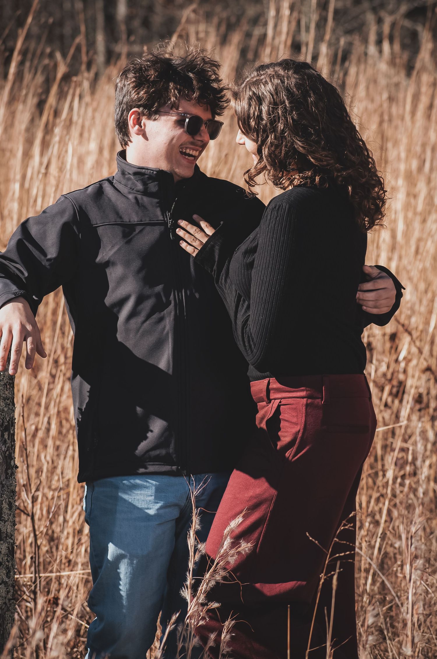 A couple embraces in a field of tall dried grass during golden hour with warm sunlight filtering through.