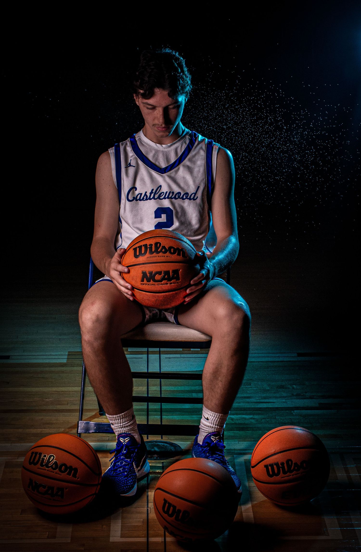 Athlete in white jersey poses with NCAA basketball surrounded by other basketballs.