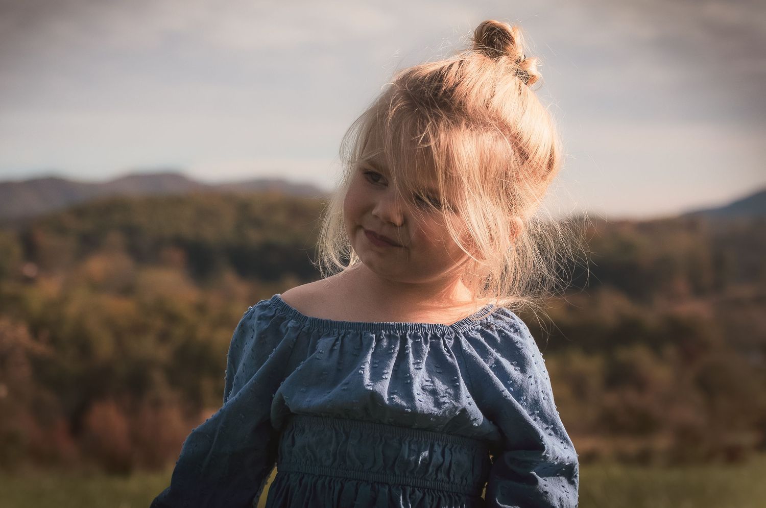 Child in blue dress smiling outdoors with mountains in background.