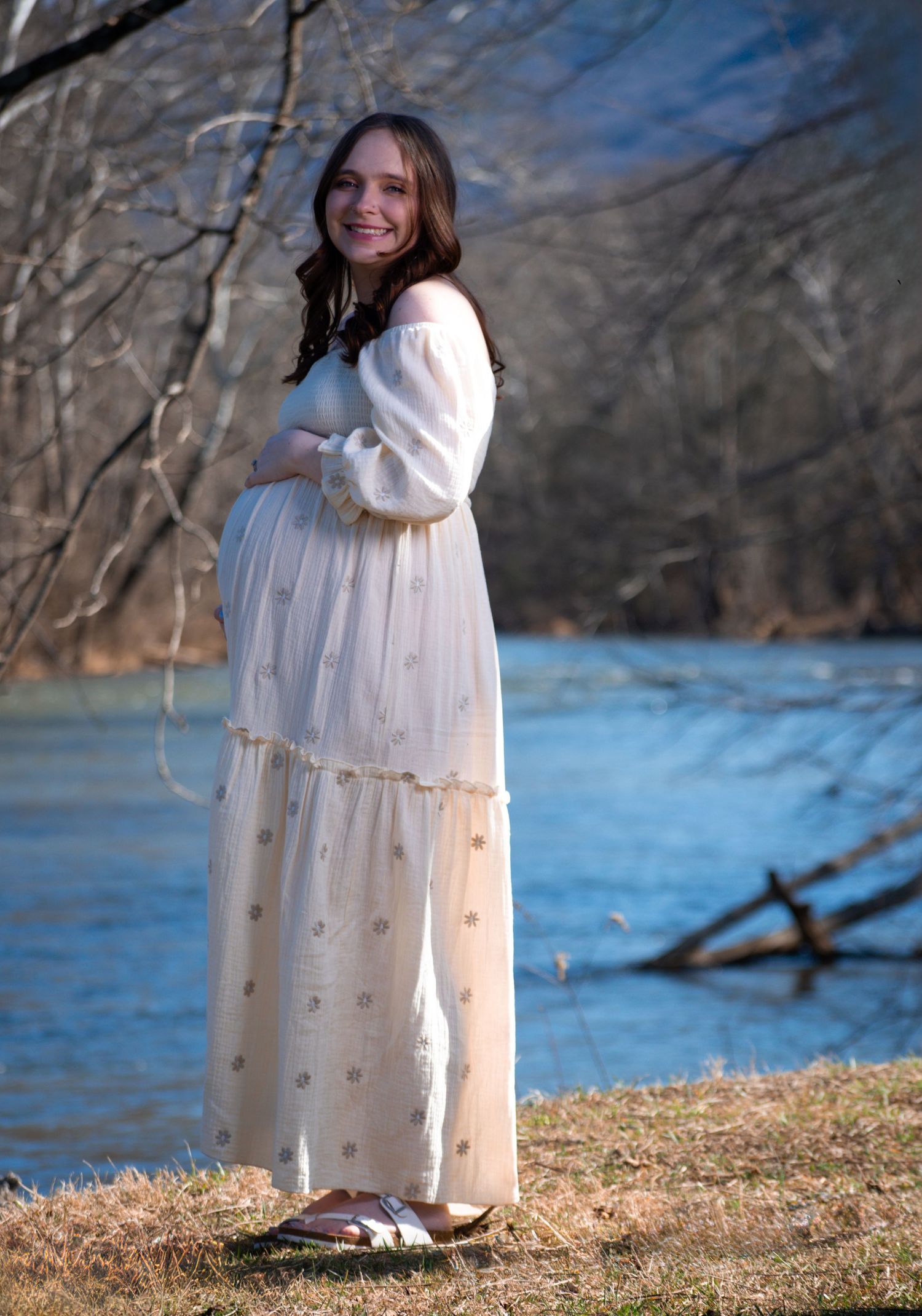 A person in a white flowing dress and sweater stands by a lake on a winter day with bare trees in the background.