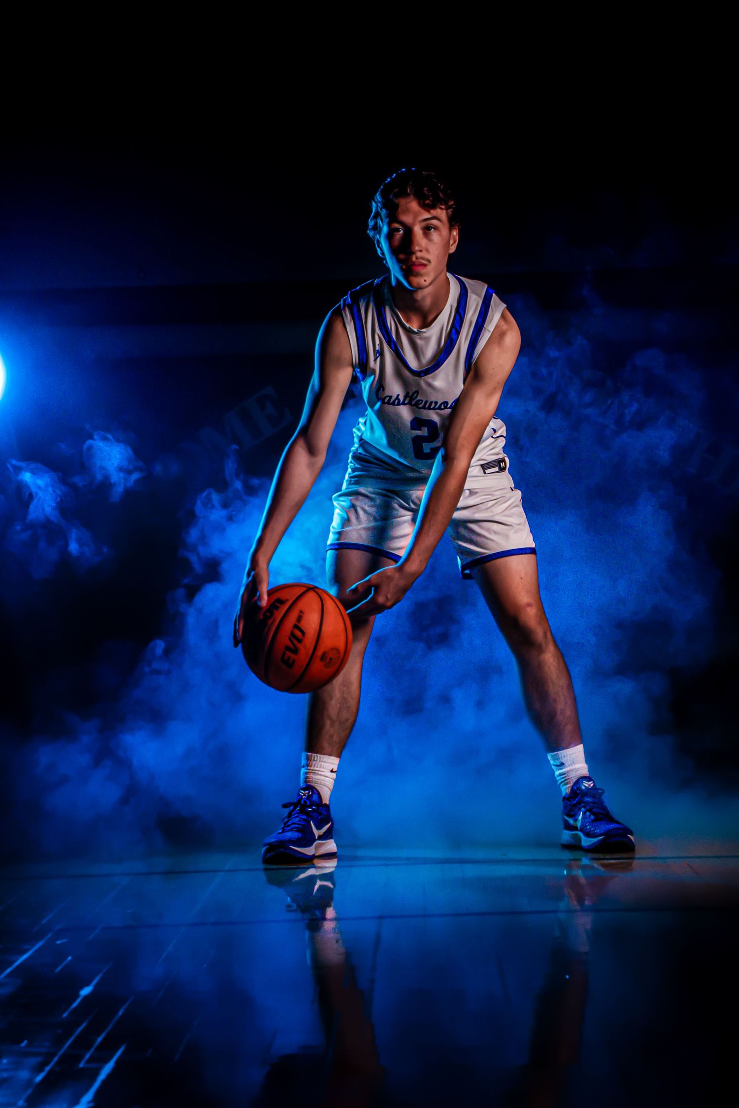 Side view of basketball player dribbling on glossy court with dramatic blue lighting and fog.