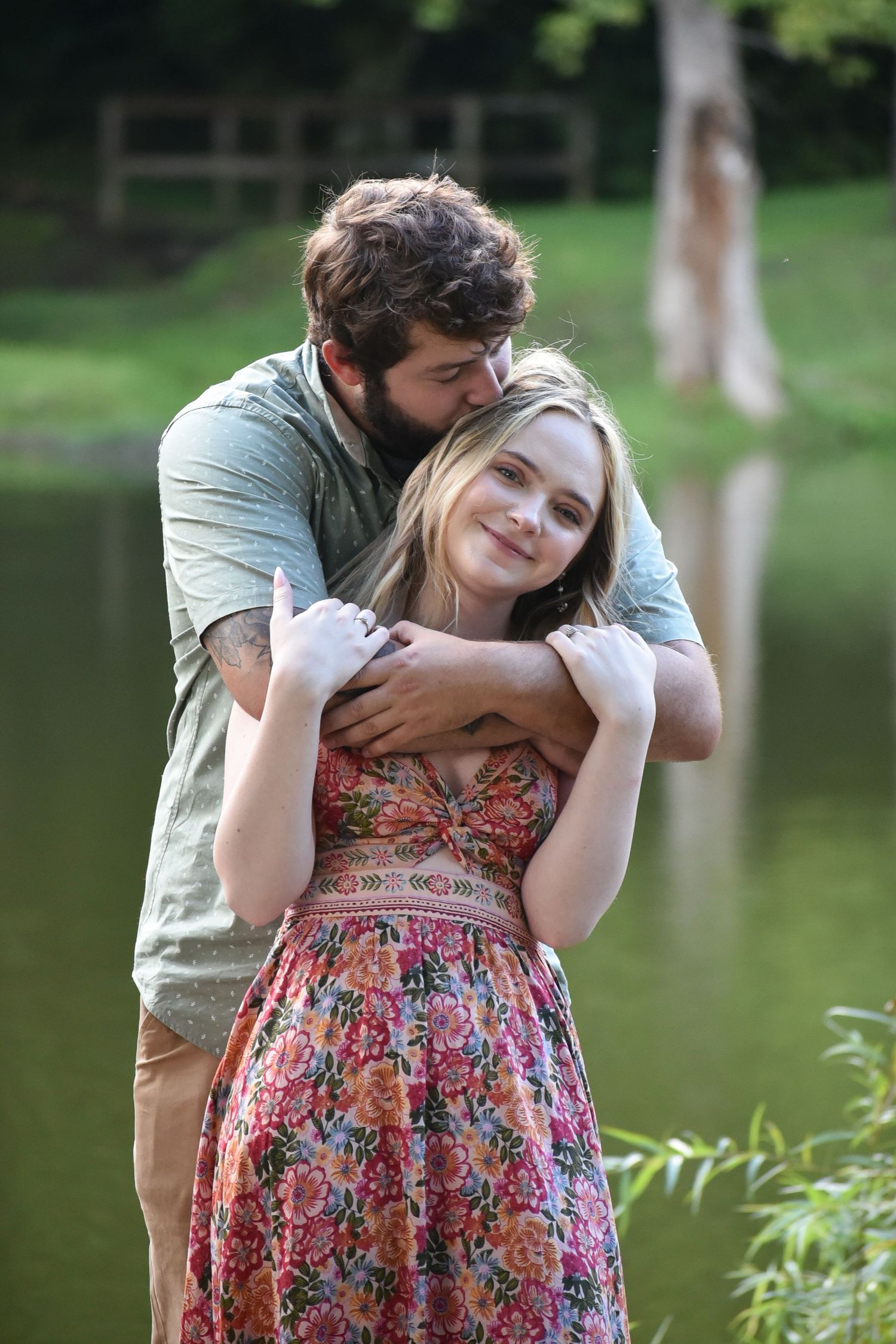 A couple embraces by a lake, with the person in a floral dress and the other in casual attire.