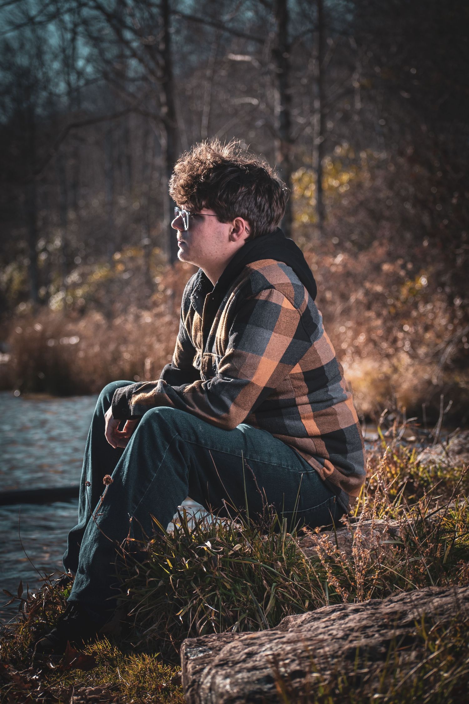 Person in plaid shirt and glasses sitting thoughtfully by water's edge during autumn.