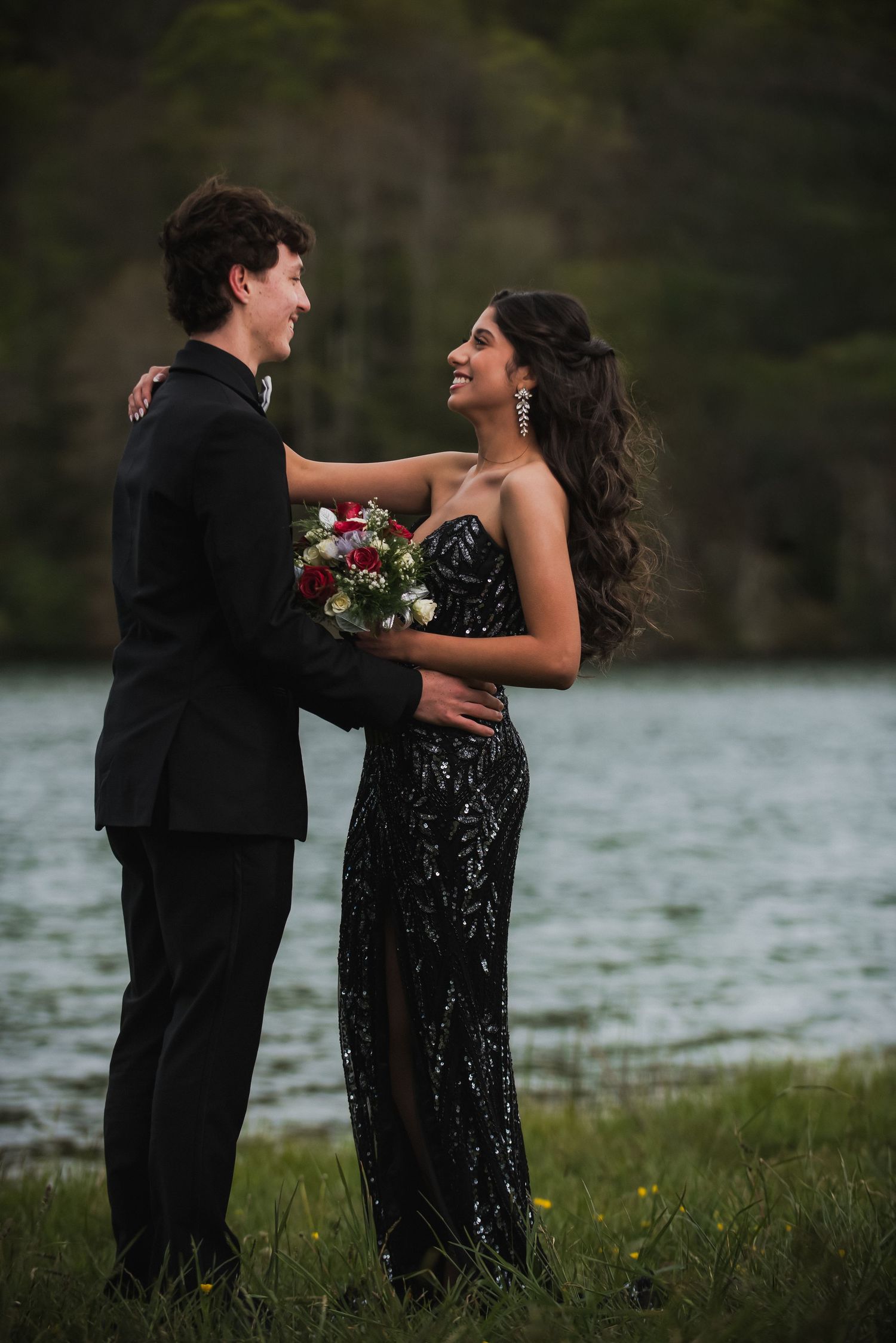 A couple in matching black formal wear poses by a lake, she holds a red and white bouquet at prom.