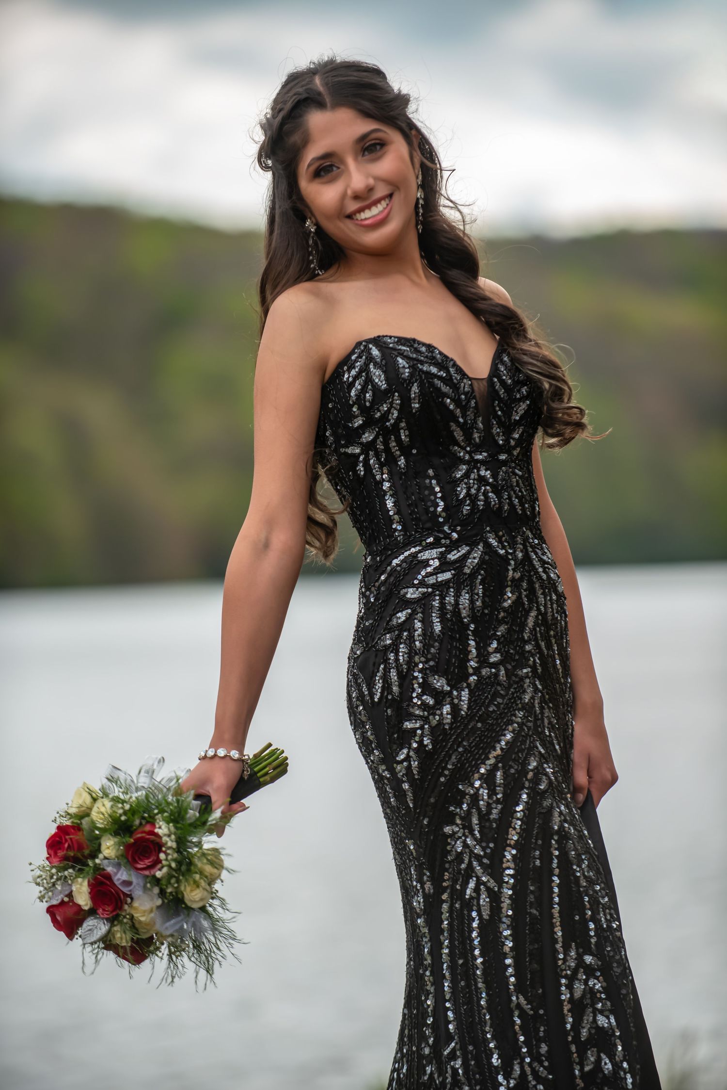 Smiling young woman in black sequin strapless gown holding floral bouquet near a lake with green hills.