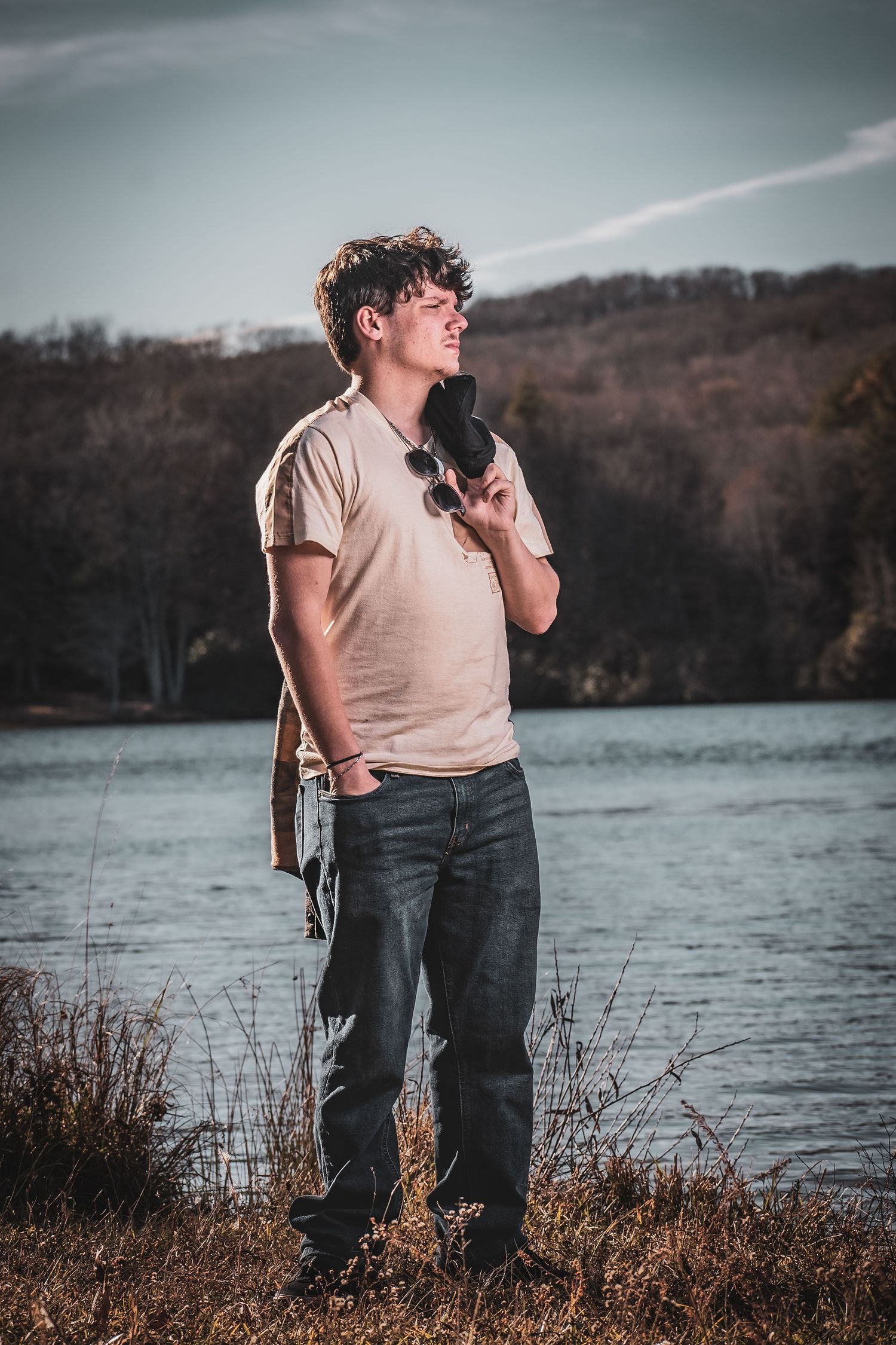 A person in a beige t-shirt and jeans stands by a lake on an autumn day with moody lighting and natural scenery.