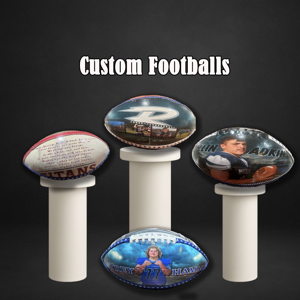 Custom football displays on white pedestals showcasing personalized designs and autographs against dark background.
