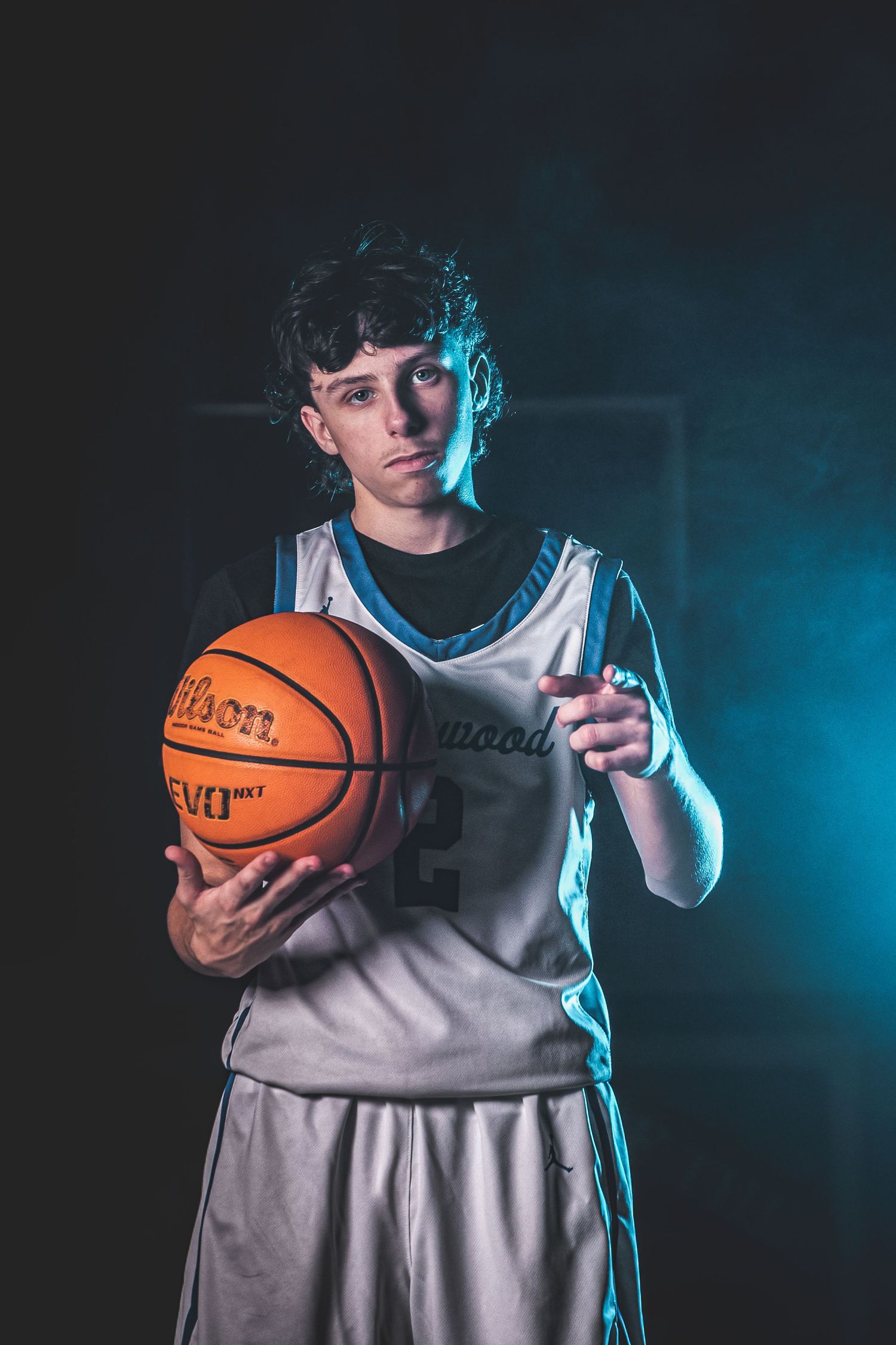 Basketball player in gray jersey holds orange ball against dramatic blue lighting in moody studio portrait.