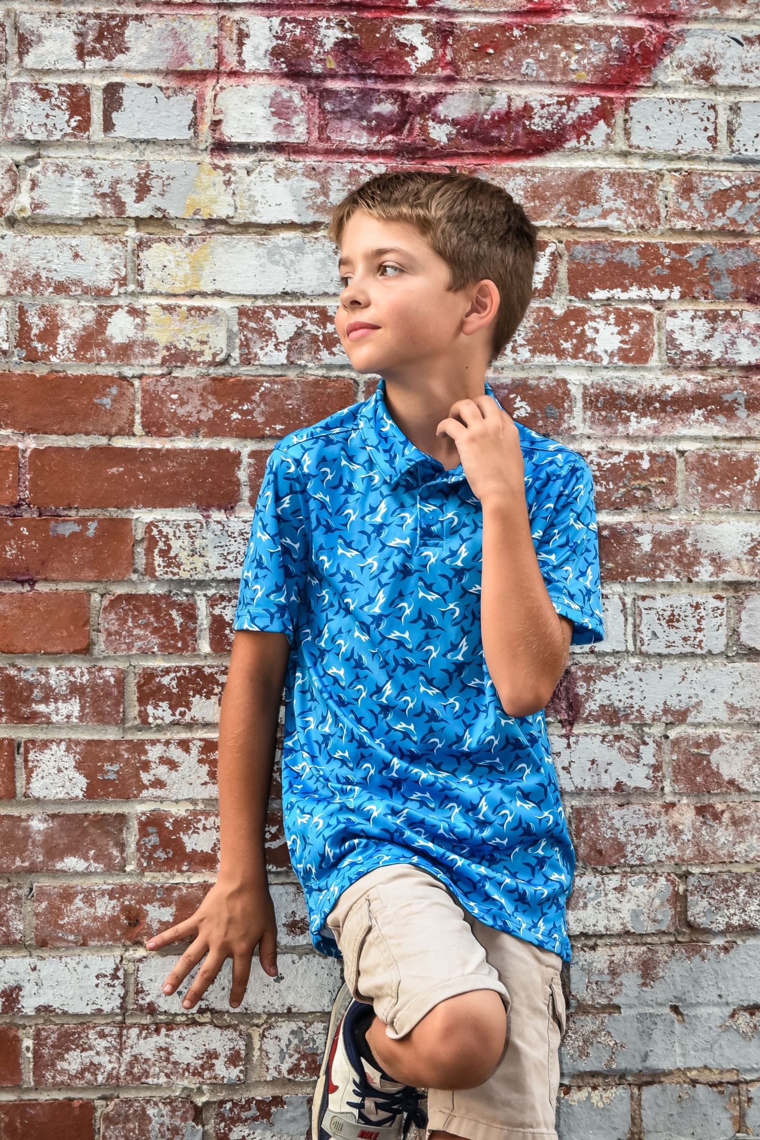 Young person in bright blue patterned shirt poses against weathered brick wall.