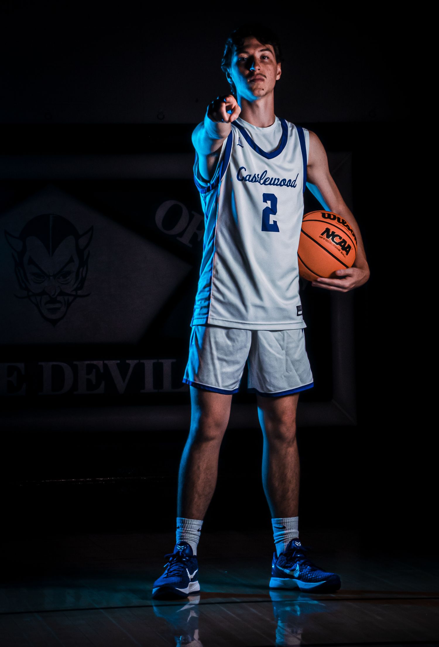 Basketball player in white uniform stands ready with ball against dark background with blue accent lighting.