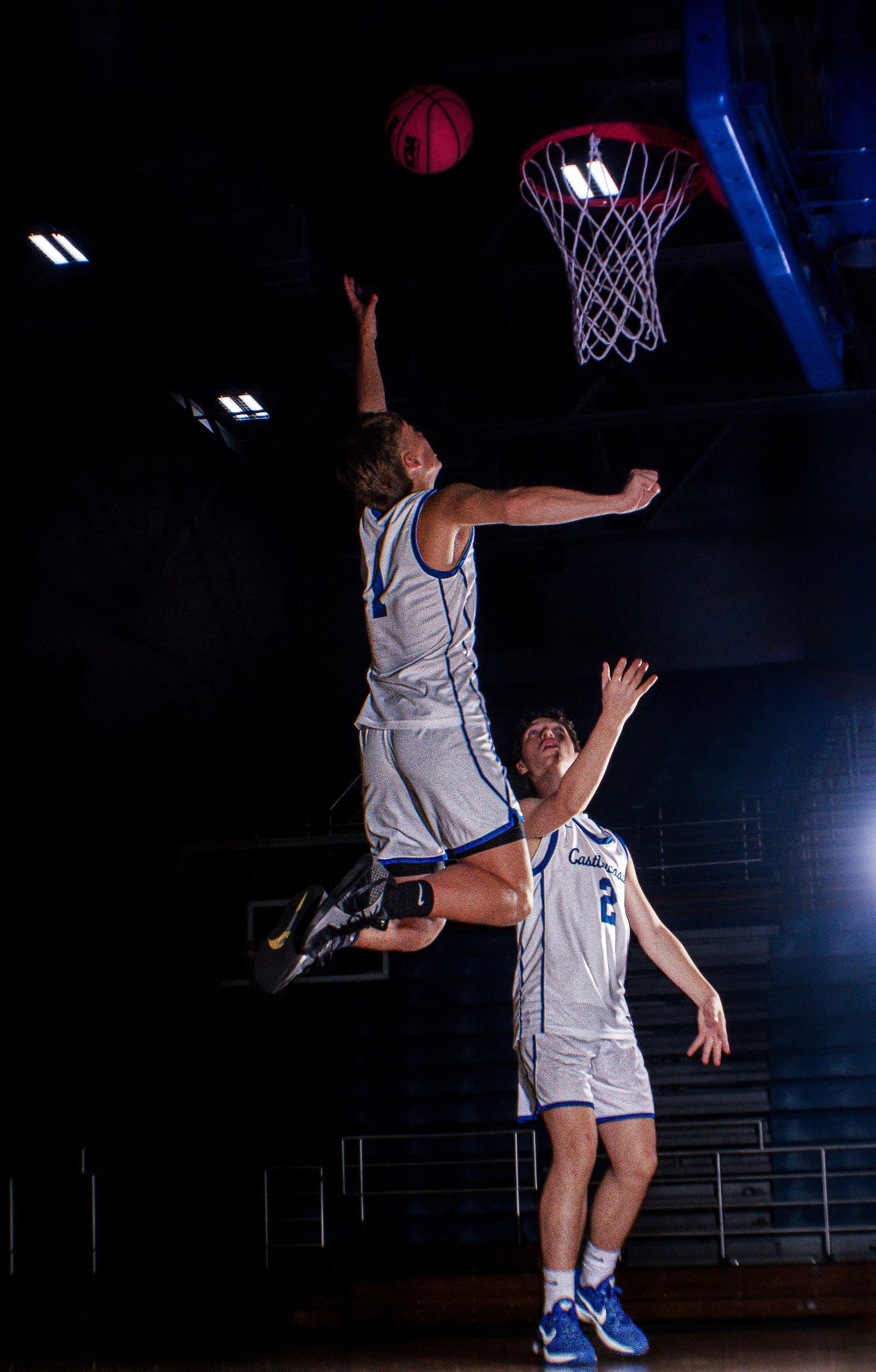 Basketball players coordinate for rebound under illuminated hoop.