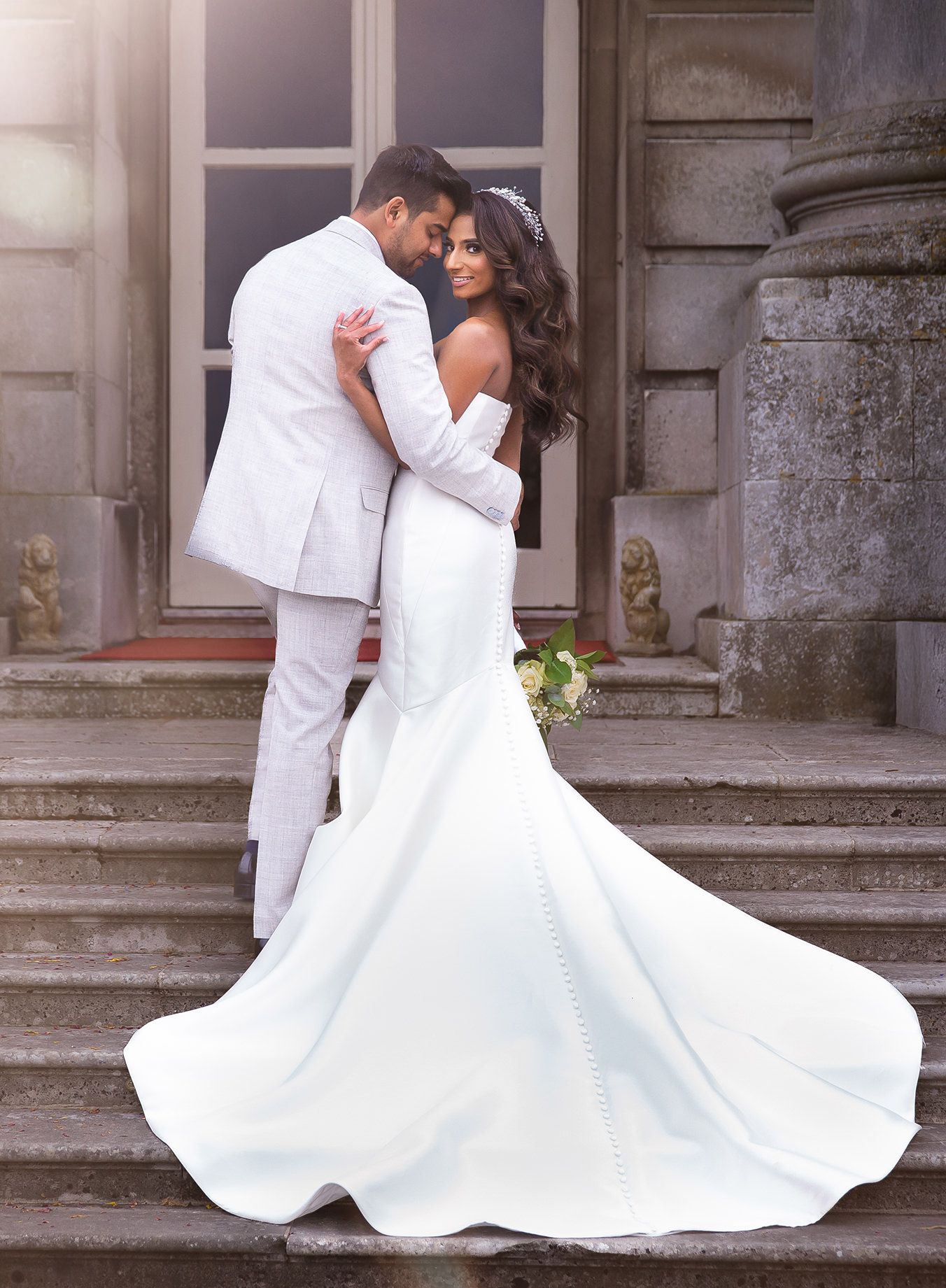 Elegant couple in white wedding attire embracing on stone steps of historic building.