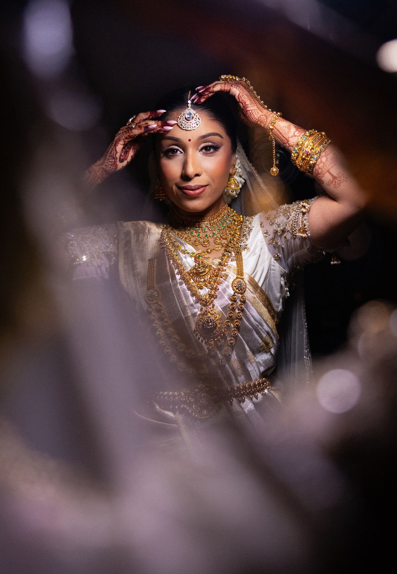 Traditional Indian bride wearing ornate gold jewelry and white attire poses elegantly in dramatic lighting.