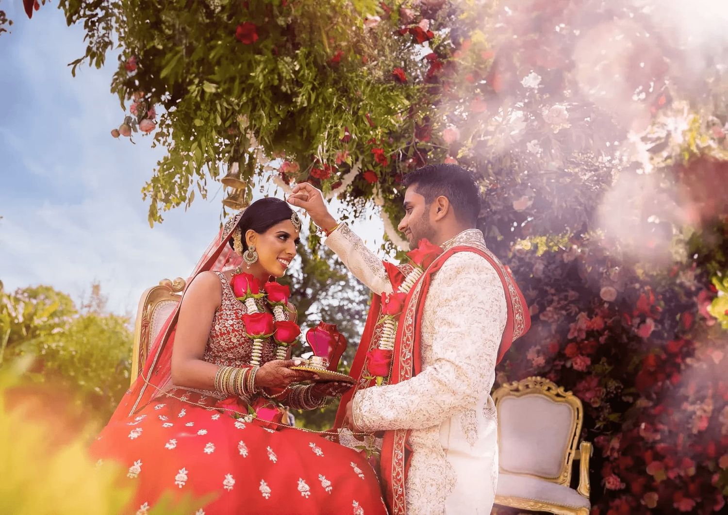 A couple in red and white traditional attire poses outdoors during a romantic photoshoot with sunlit trees in background.