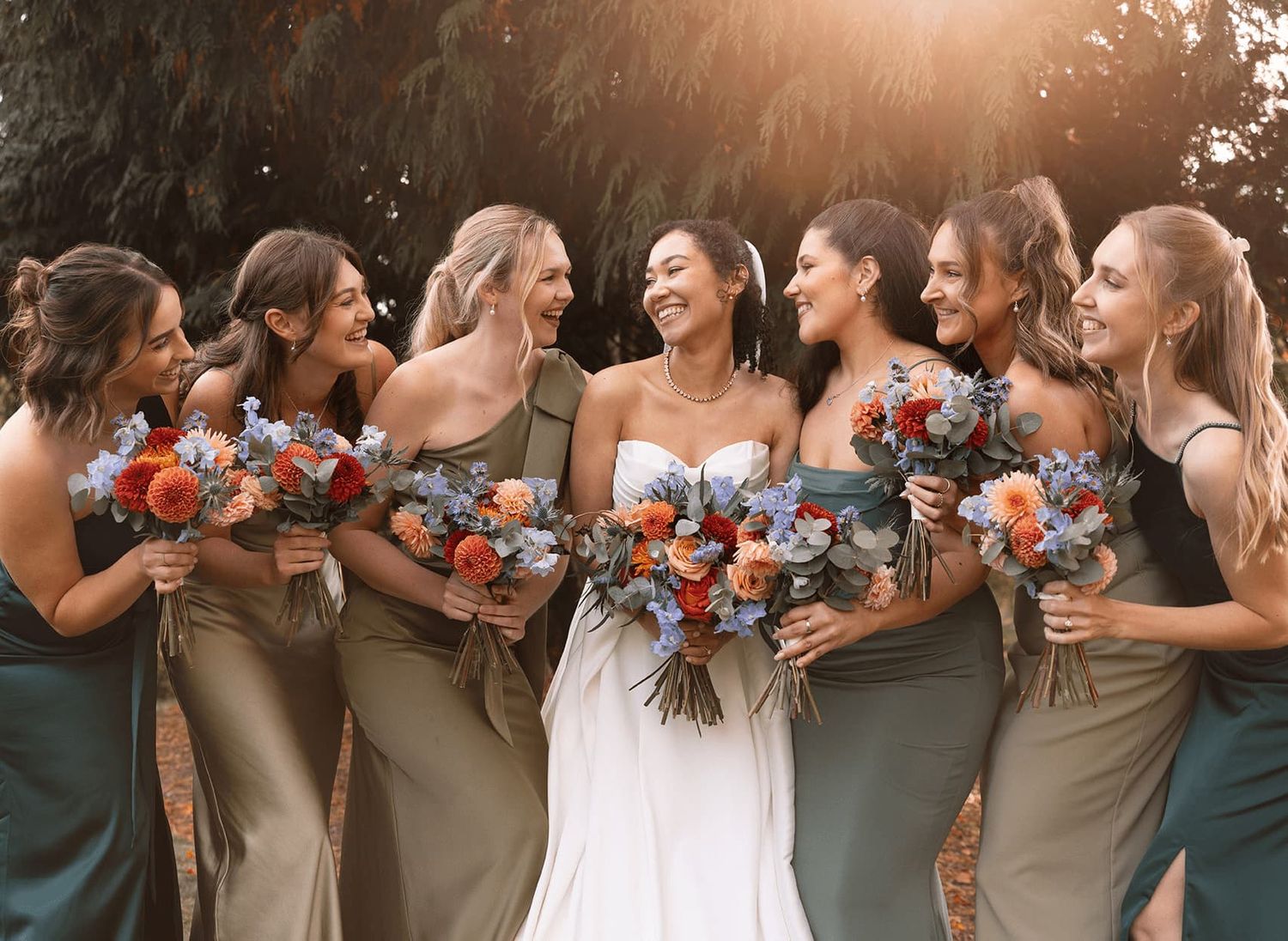 Wedding party members in teal and tan dresses stand together holding vibrant blue and coral bouquets.