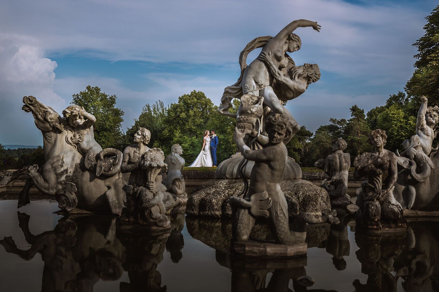 Stone statues of cherubs and angels decorate an ornate classical fountain against a blue sky.