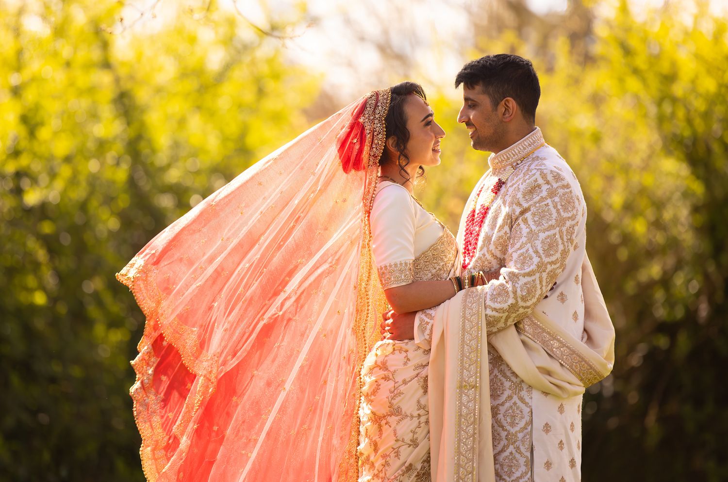 Couple in traditional Indian wedding attire embracing in front of yellow flowering field.