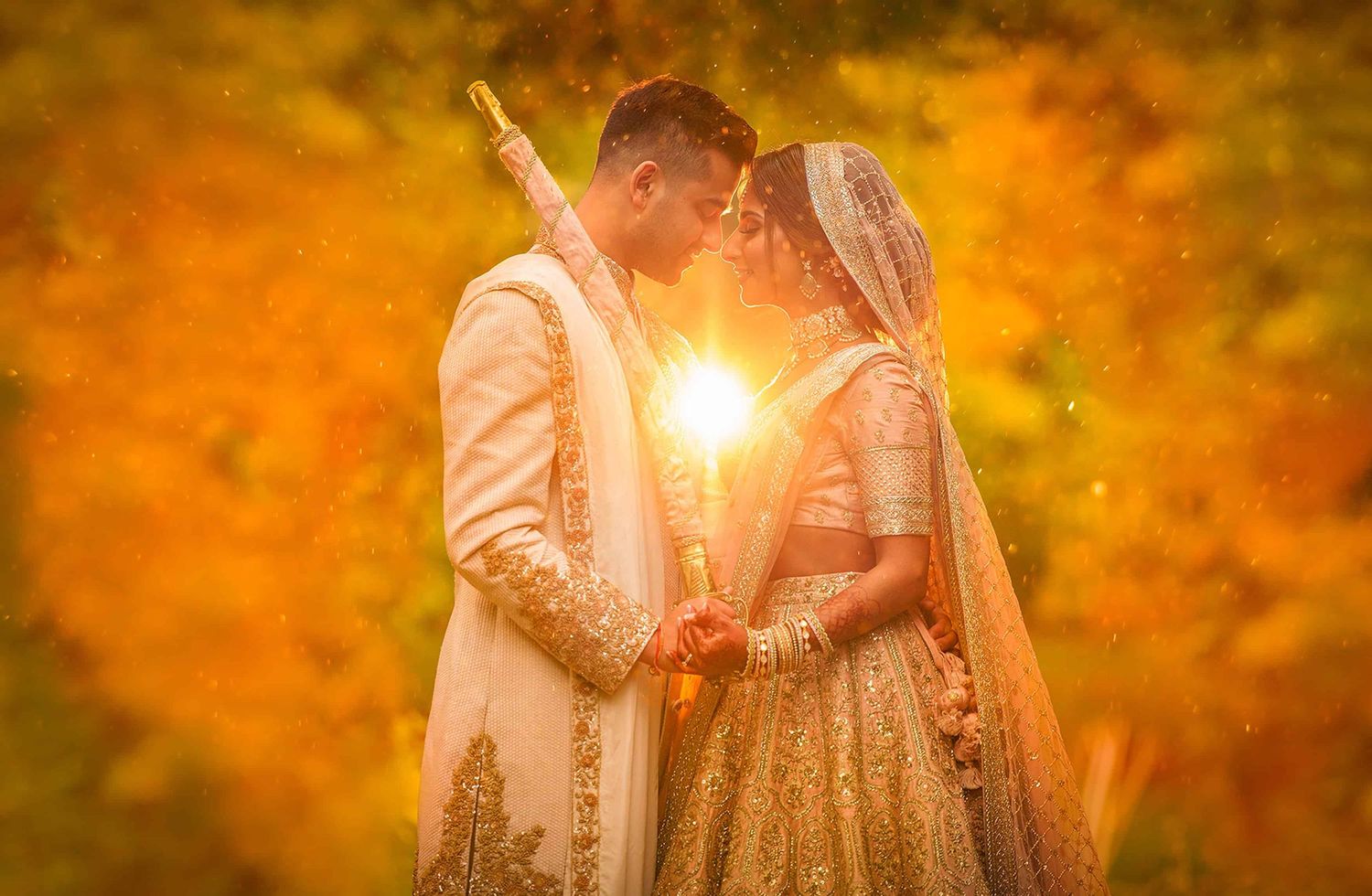 A couple in elegant wedding attire poses in a golden autumn forest with sunlight streaming through the trees.