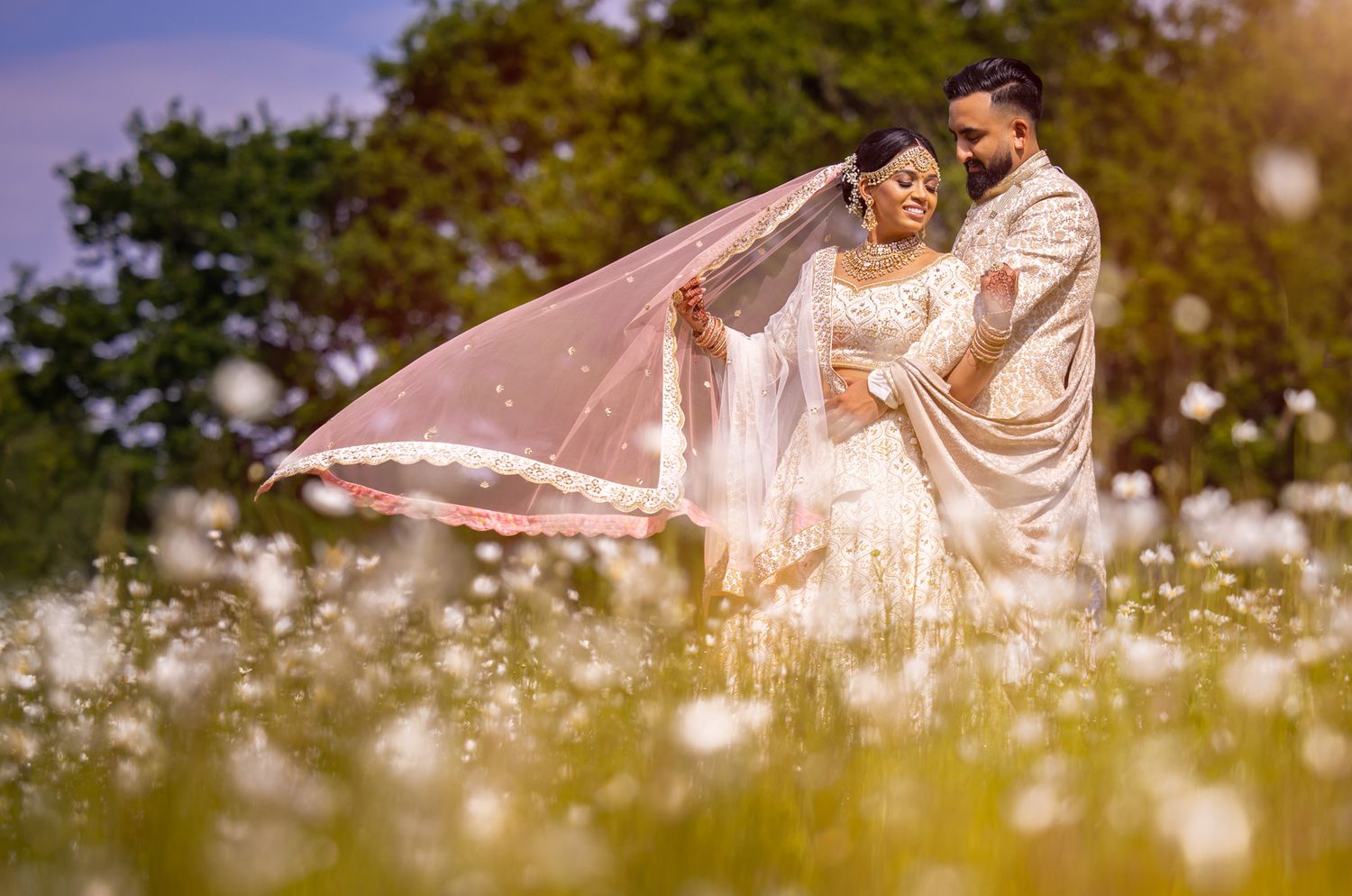 Romantic wedding couple in white attire with flowing veil standing among wildflowers.