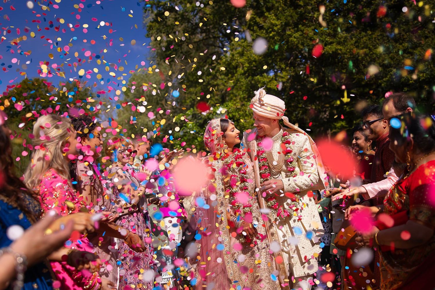 Colorful confetti and flower petals shower over a traditional Indian wedding celebration in bright daylight.