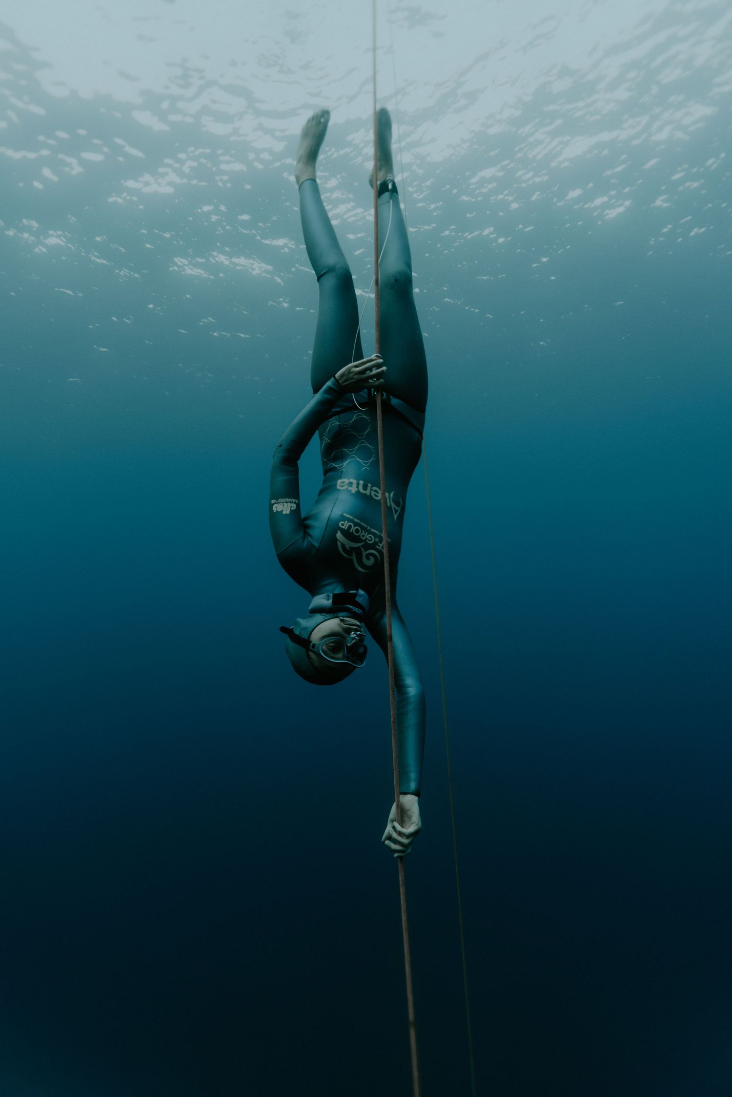 Freediver in wetsuit descending vertically along a rope in clear blue waters.
