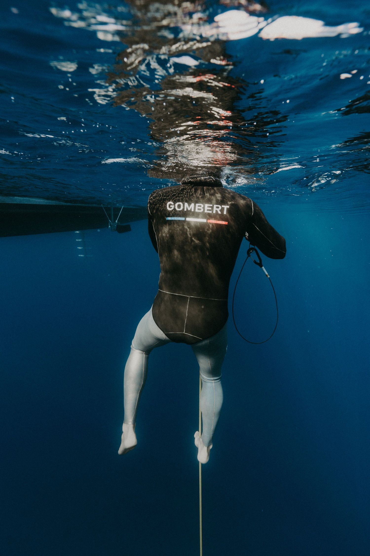 Freediver in a Gombert wetsuit descending underwater in clear blue ocean, viewed from below the surface.