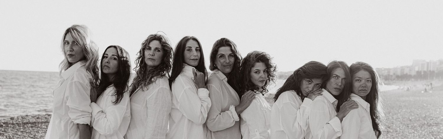 Black and white photo of nine women in white shirts posing together outdoors near a beach.
