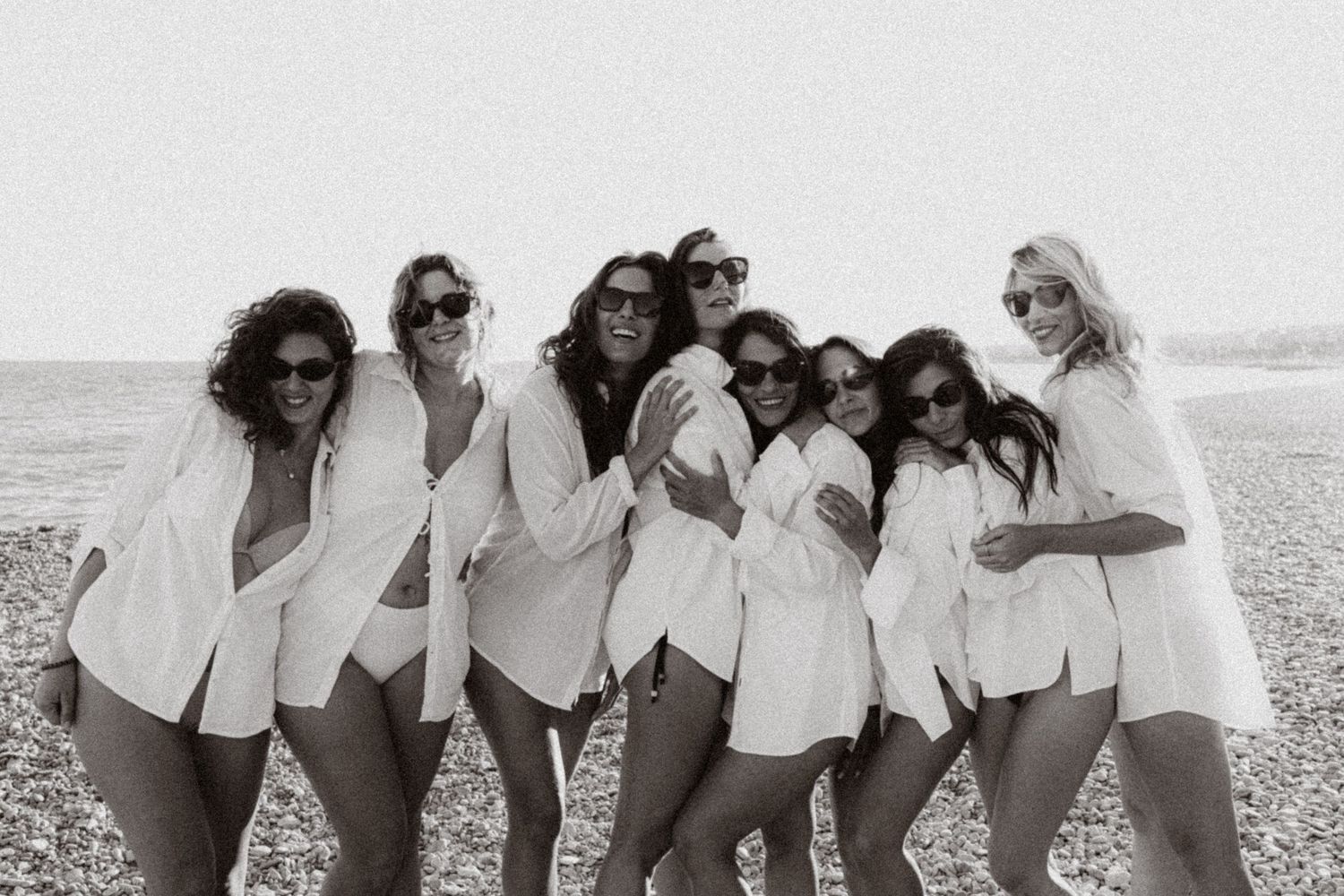 Group of women in white shirts and sunglasses posing together on a beach in a black and white photo.