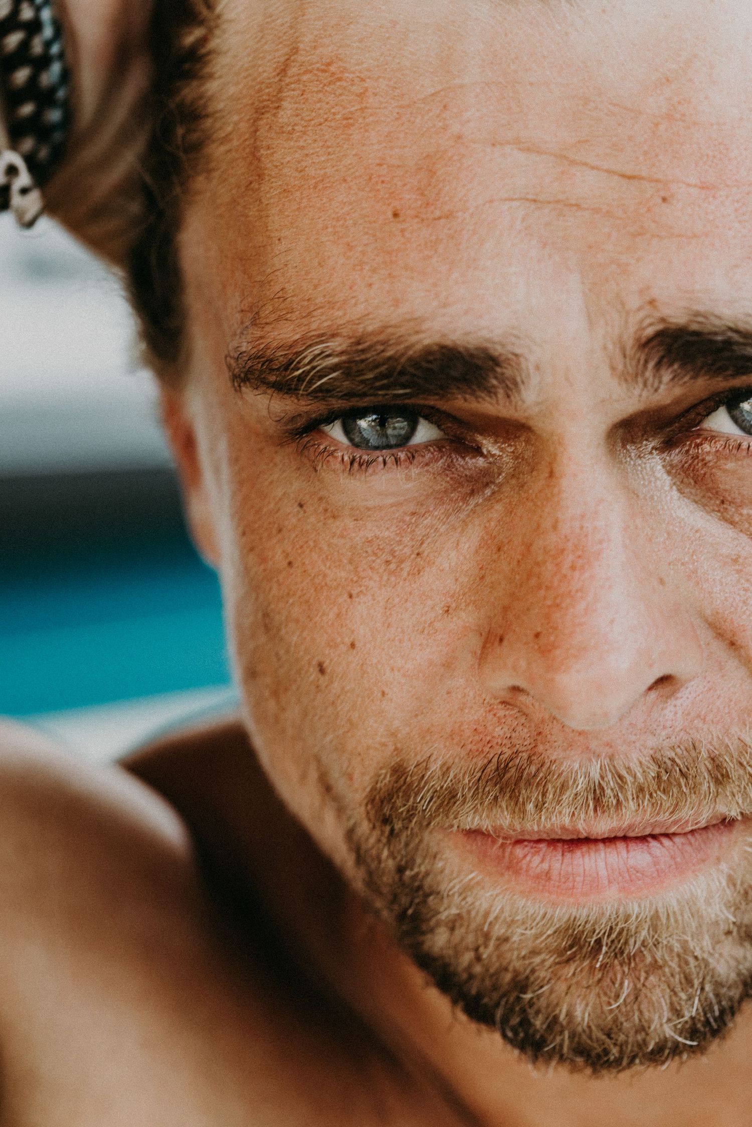 Close-up portrait of a man with blue-green eyes, stubble beard, and freckles outdoors.