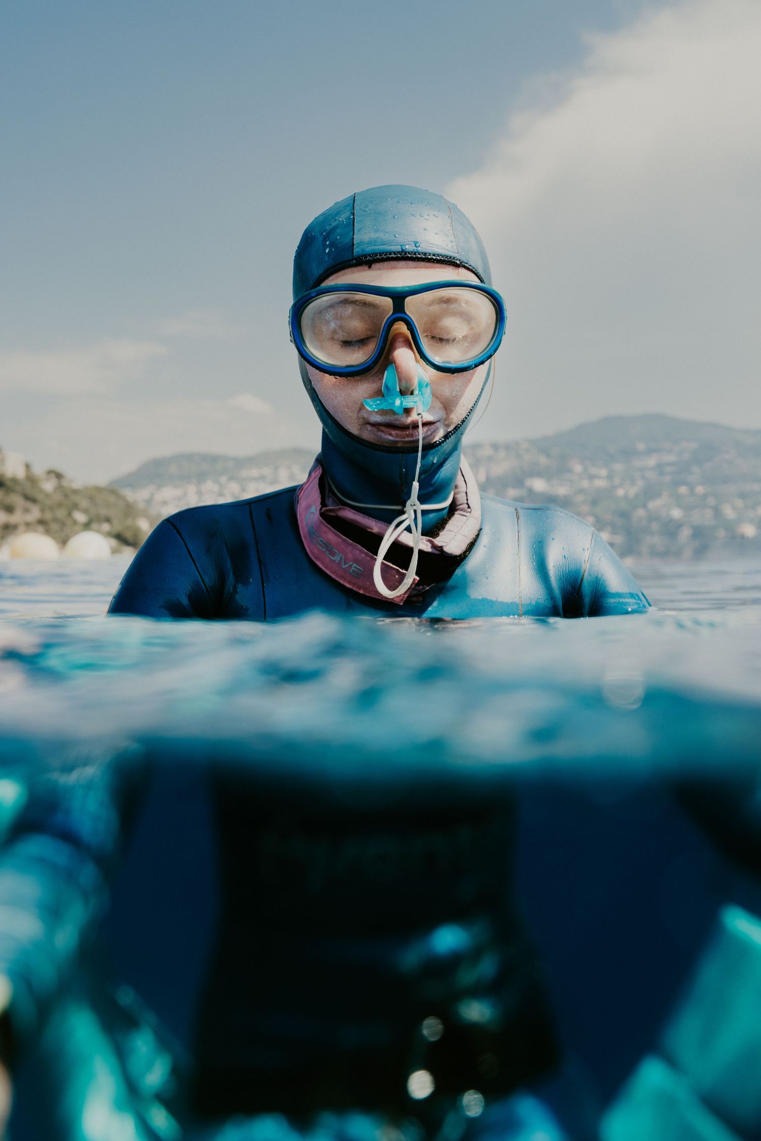 Freediver in wetsuit and mask floats at ocean surface with mountains in background on sunny day.