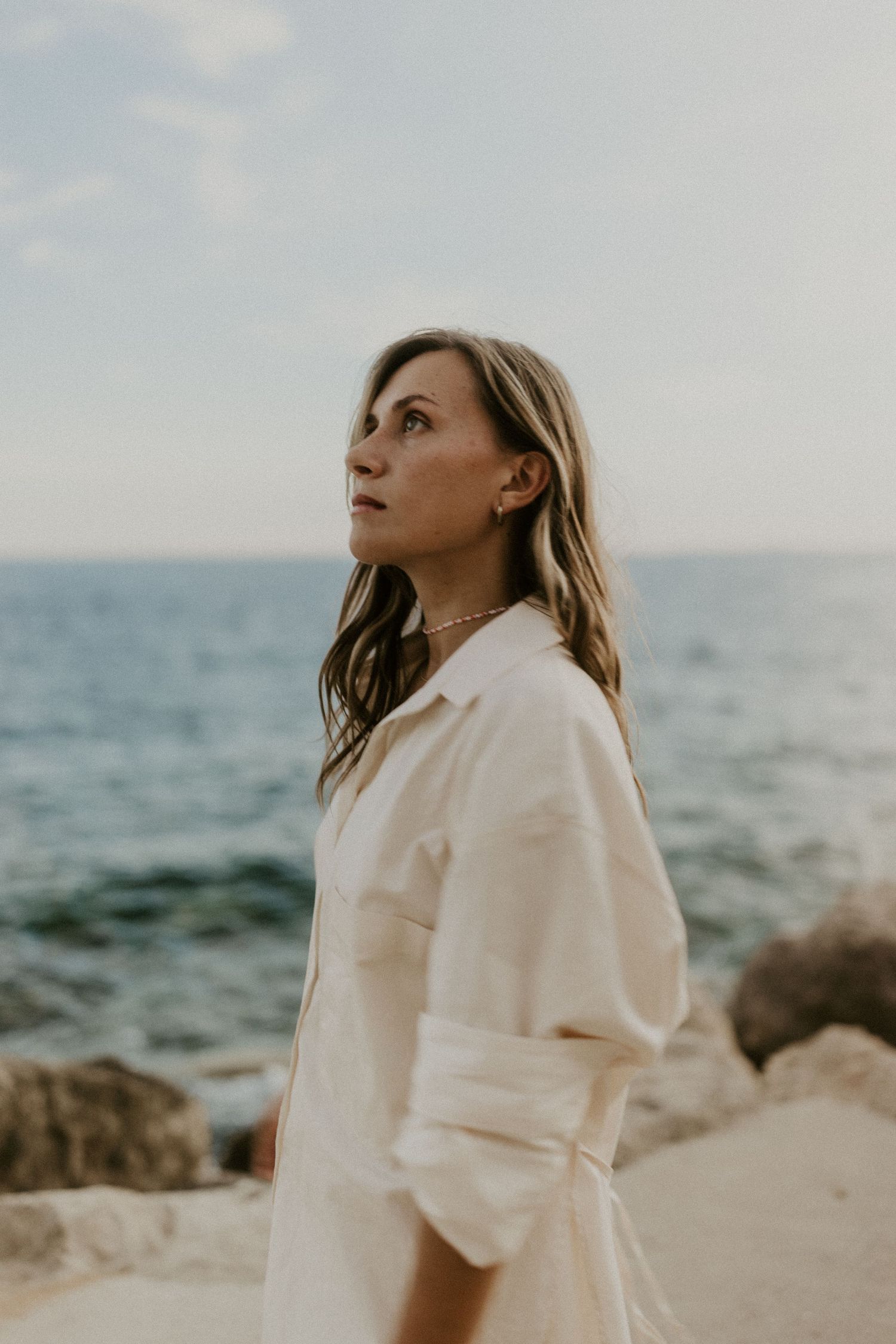 Woman in white linen shirt gazing upward by rocky ocean shore on cloudy day.