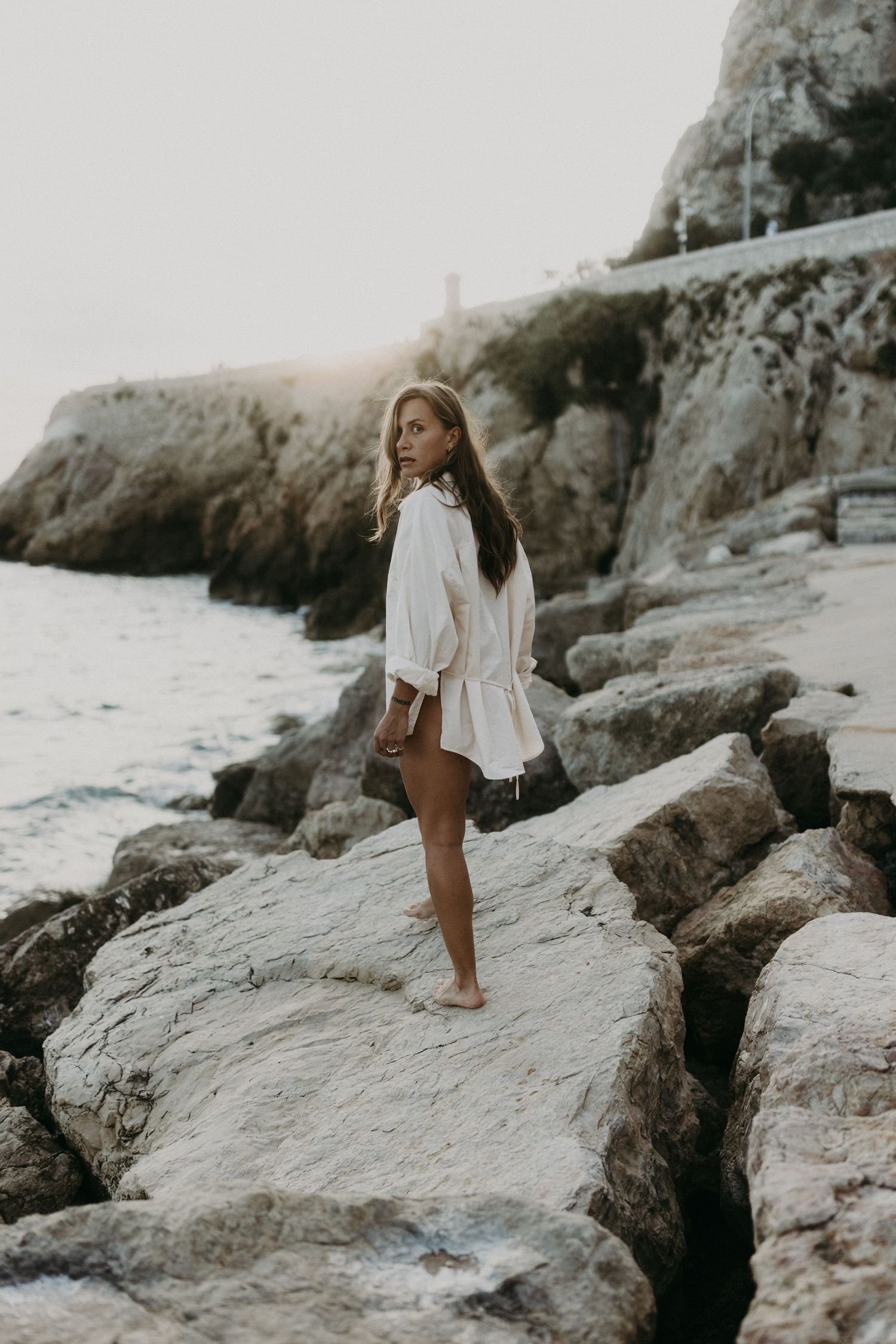 Young woman in white blouse standing barefoot on coastal rocks at sunset with cliffs in background.