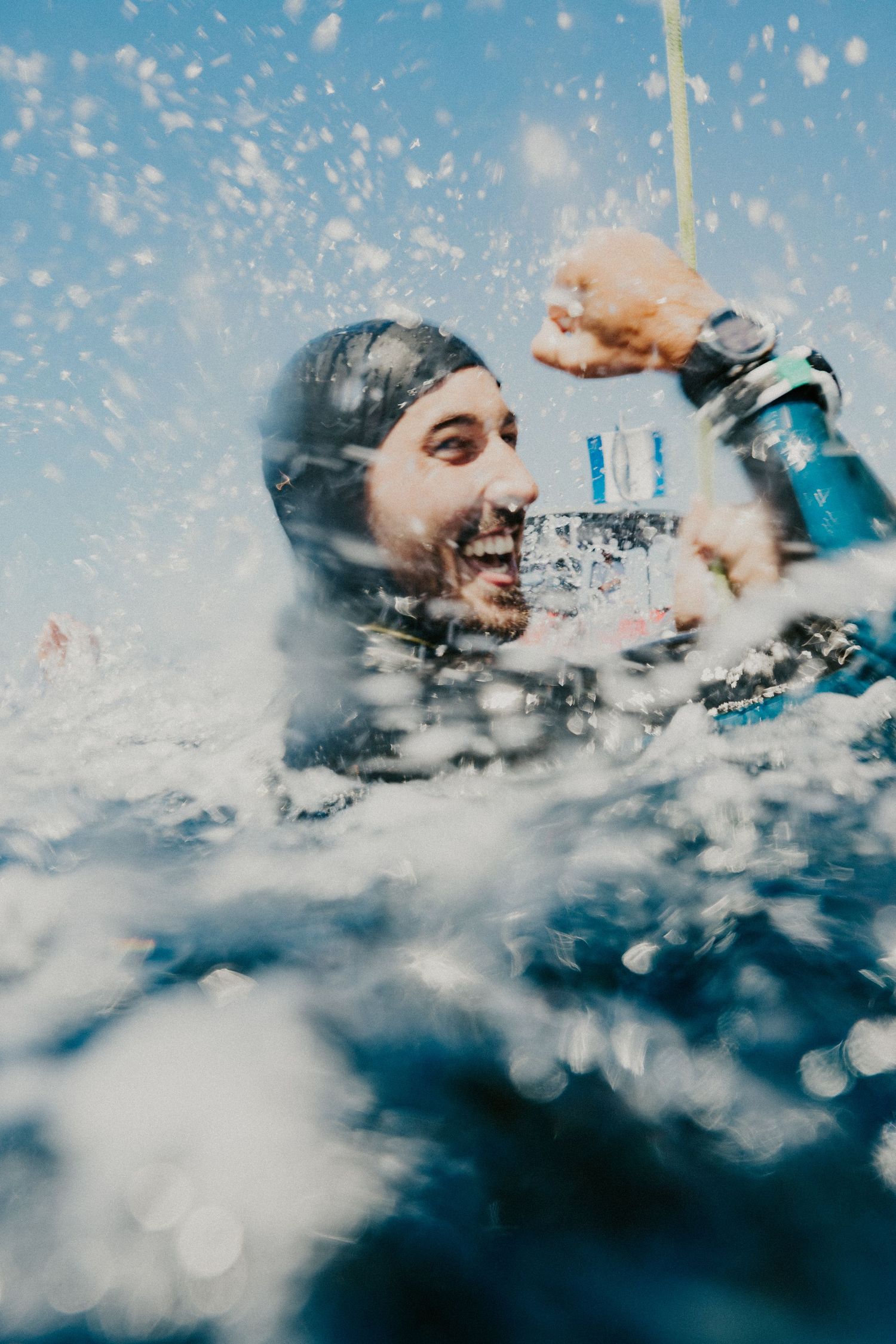Smiling scuba diver emerging from ocean waves in wetsuit and diving gear on a sunny day.