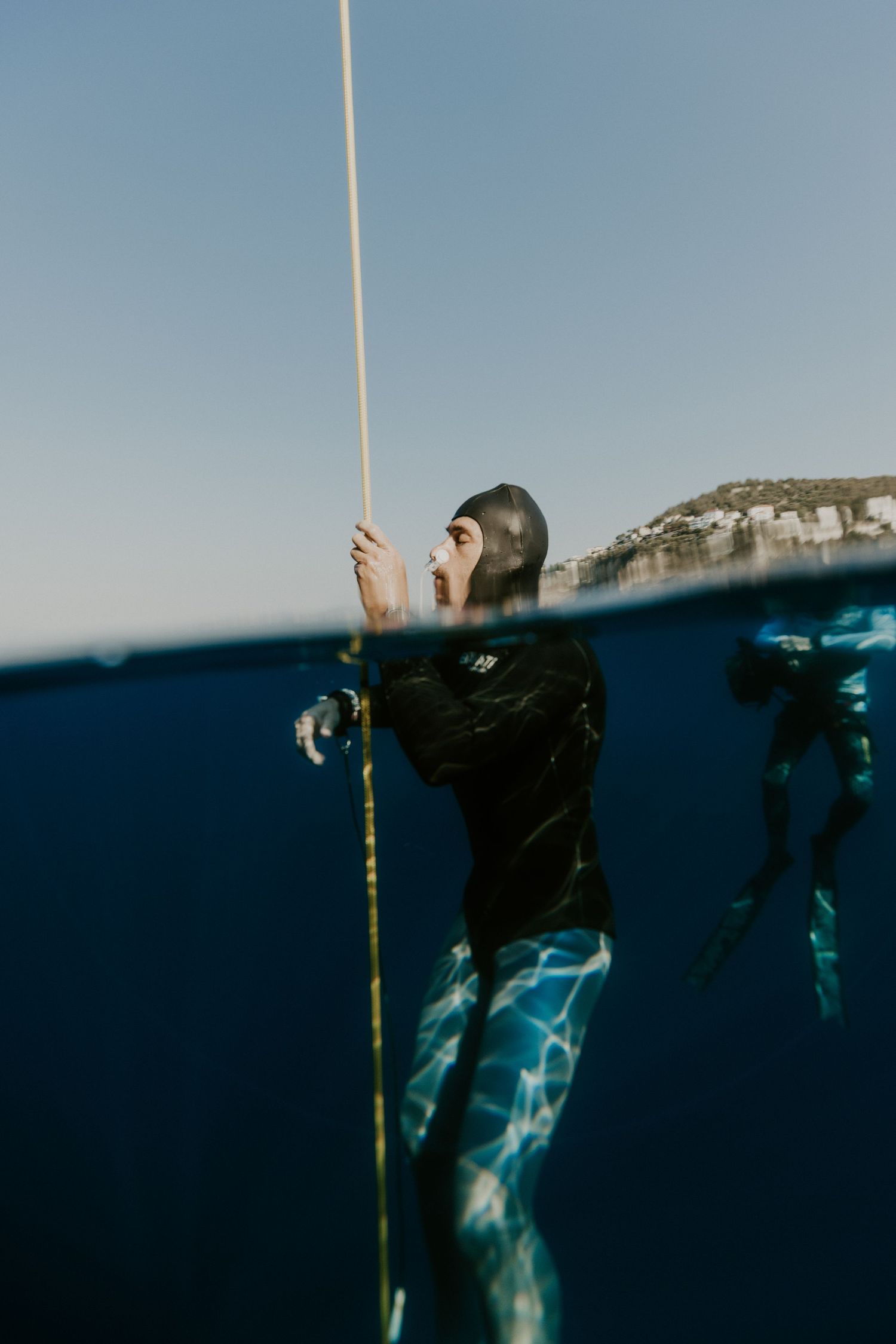 Freediver in wetsuit holding a rope at the ocean surface, with another diver visible underwater near a coastal cliff.