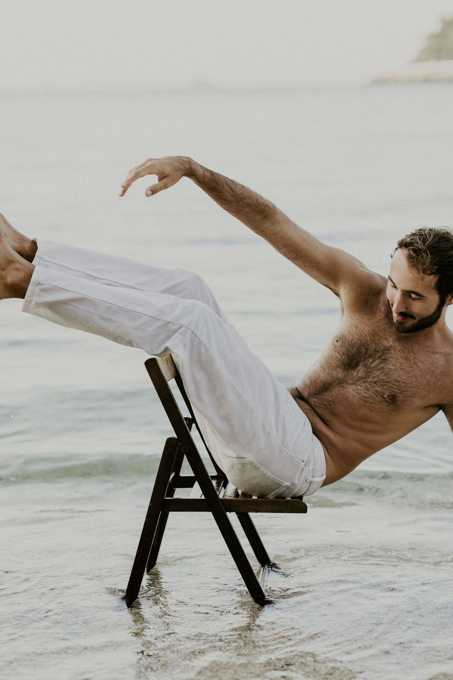 Athletic figure balancing on chair in ocean water wearing white pants in artistic pose.