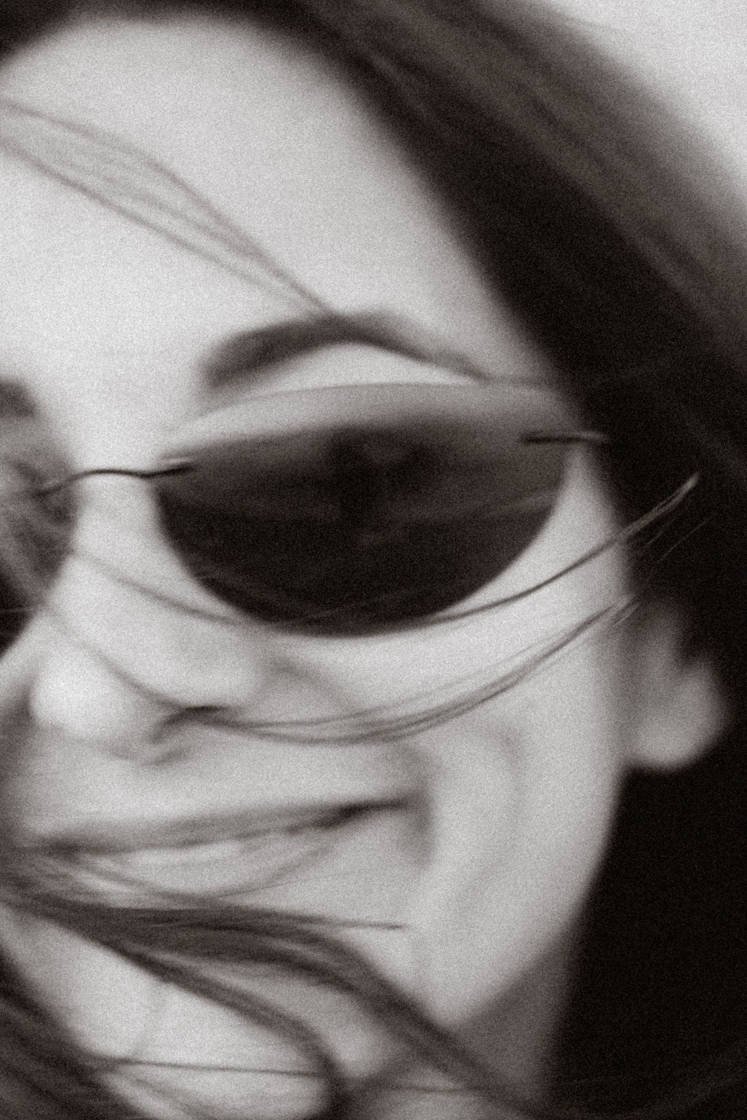 Black and white close-up of a smiling woman wearing sleek sunglasses with hair blowing across her face.
