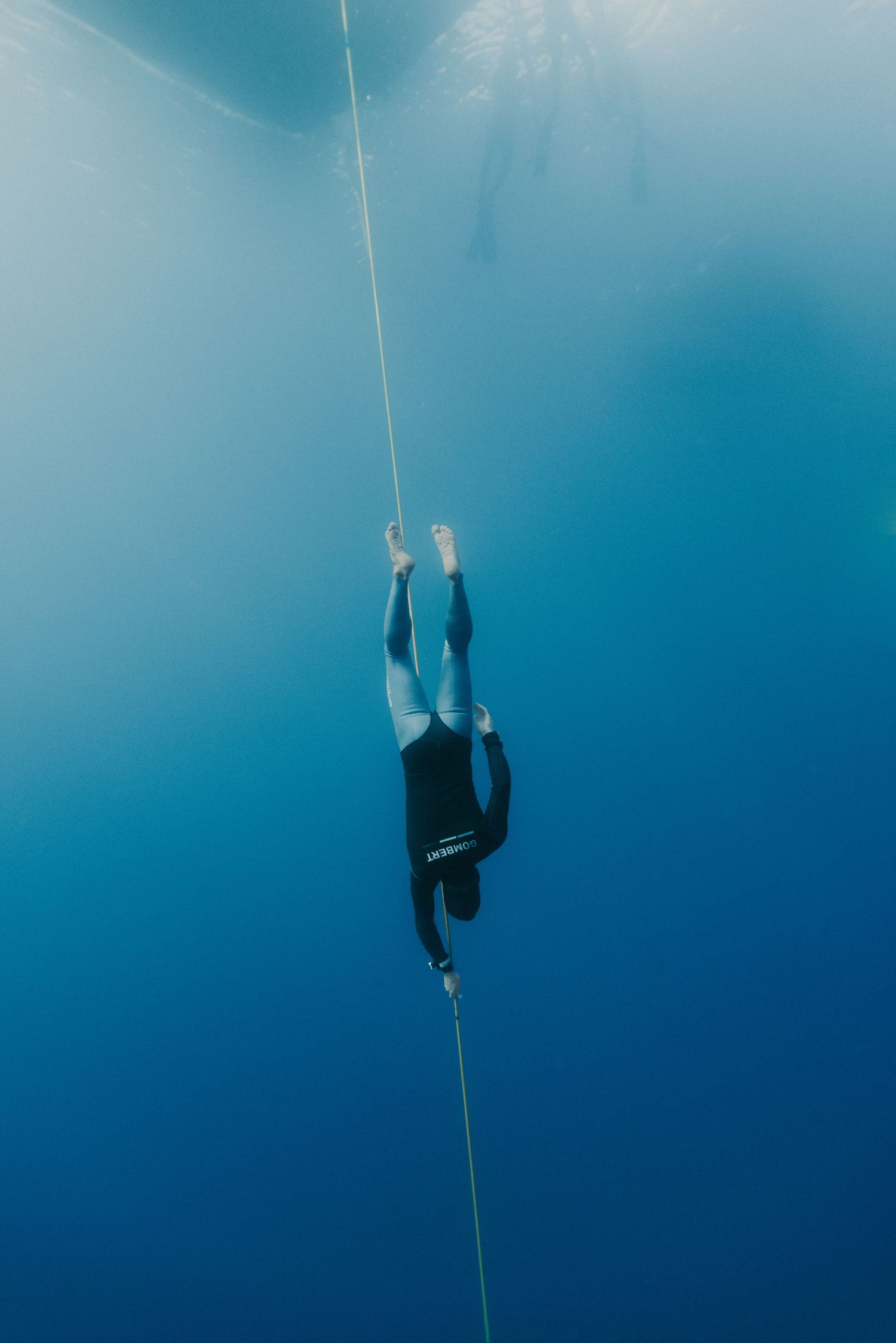 Freediver in wetsuit descends along a rope into deep blue ocean water, viewed from below with sunlight above.