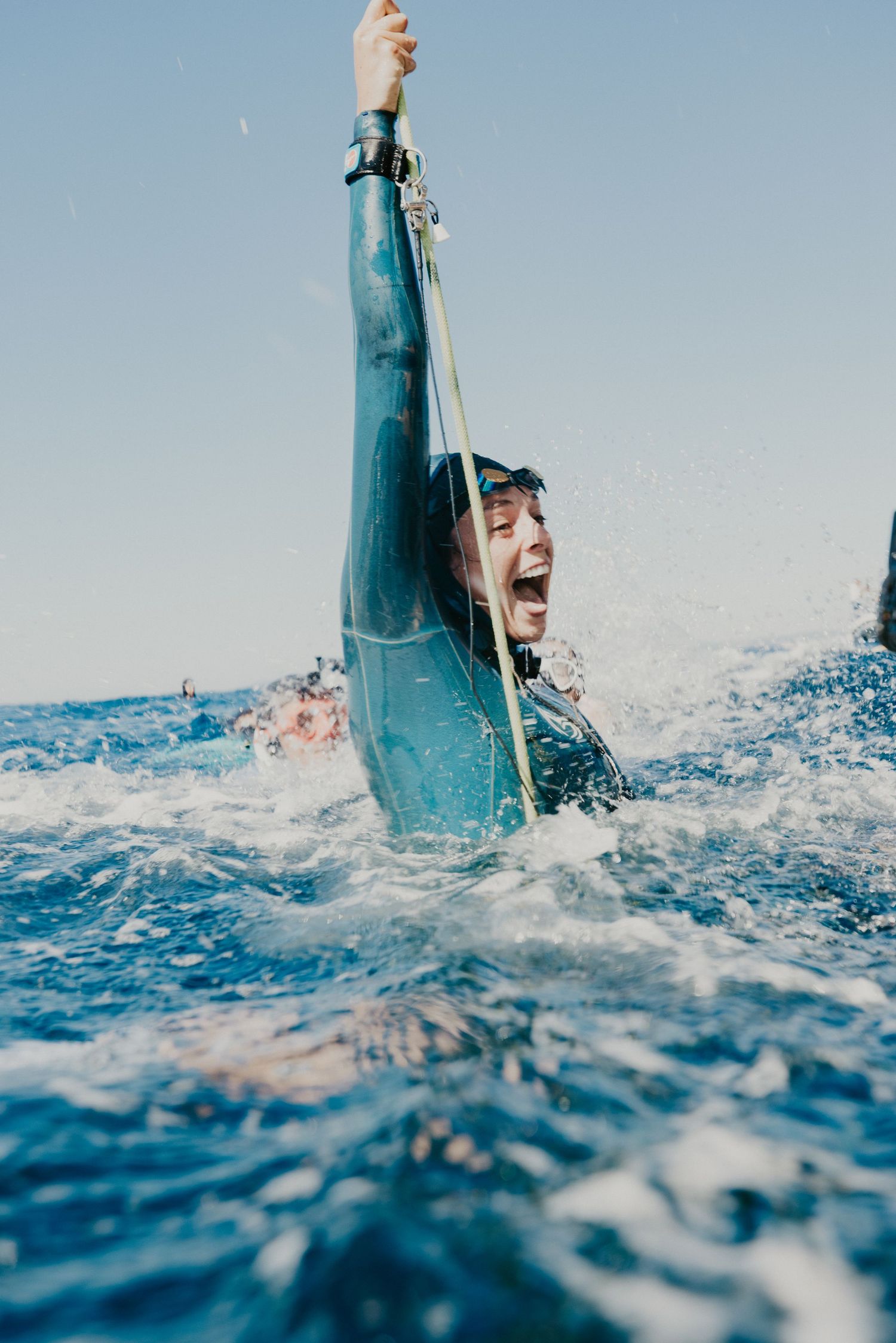 Excited freediver in wetsuit emerging from blue ocean waves, raising rope triumphantly with a joyful smile.