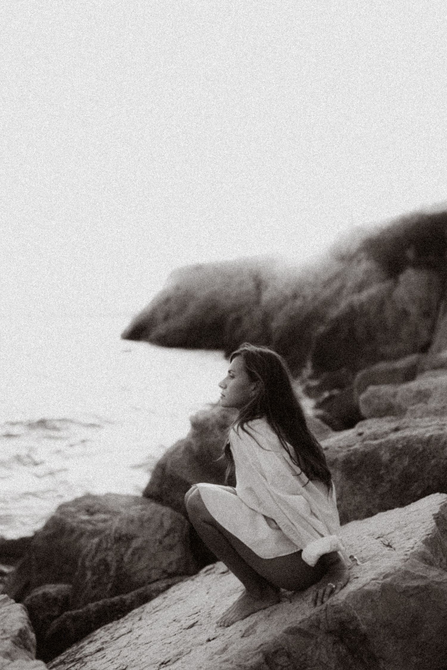 Black and white photo of a young woman sitting on rocky coastal cliffs, gazing at the ocean in a flowing white shirt.