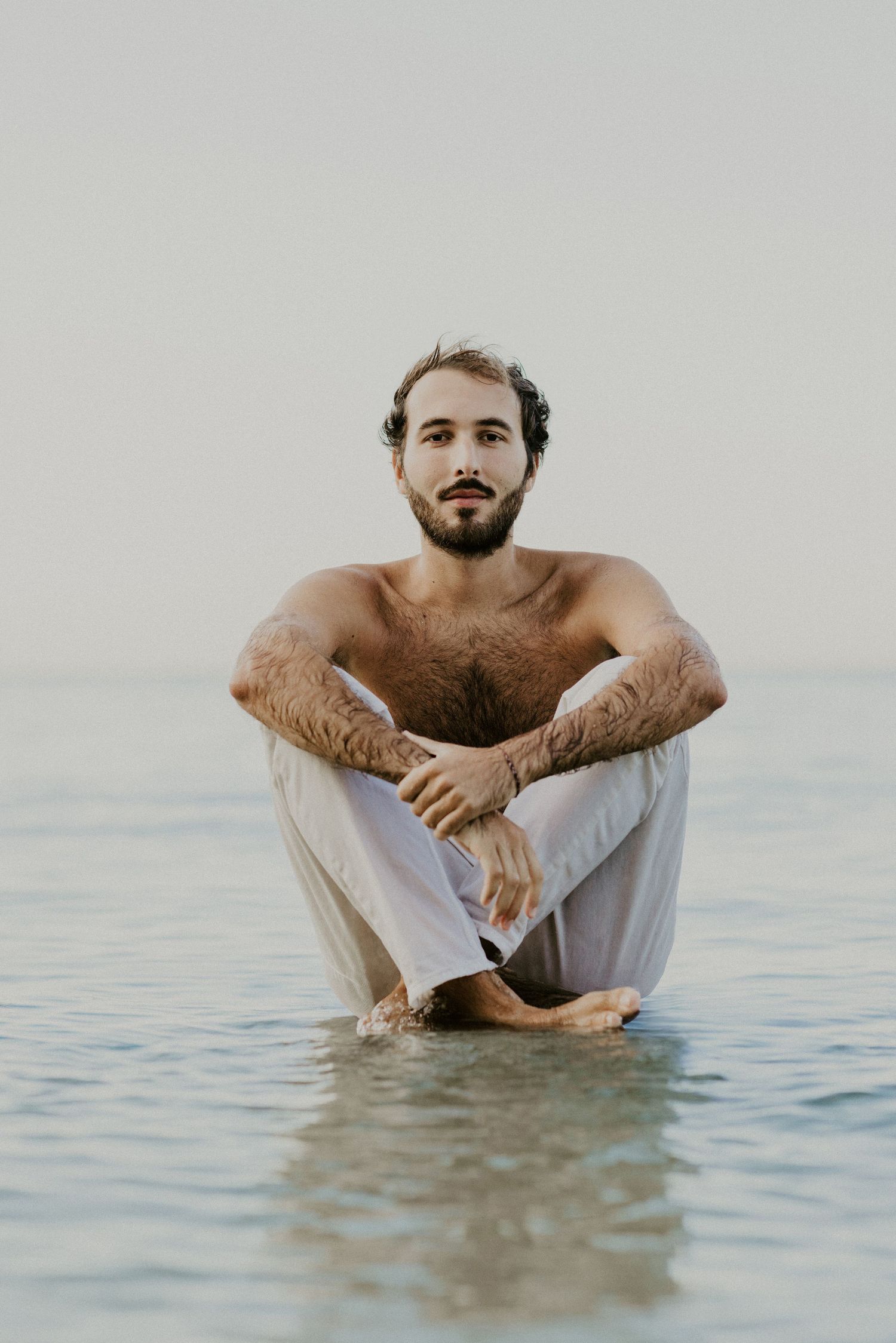 Person in white pants sitting cross-legged in calm ocean waters during a serene moment.