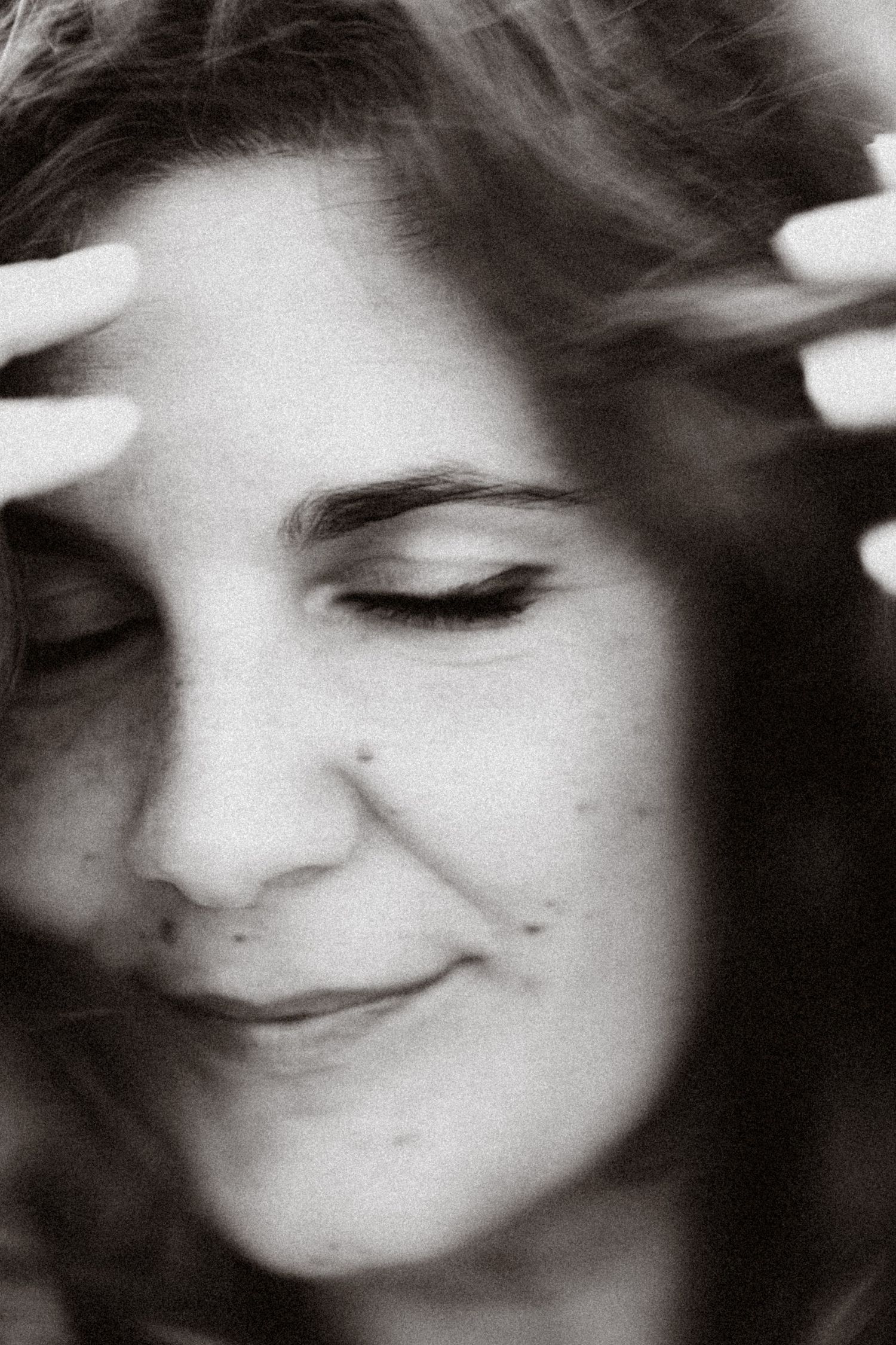 Black and white close-up portrait of a woman with eyes closed, hands in hair, smiling softly.
