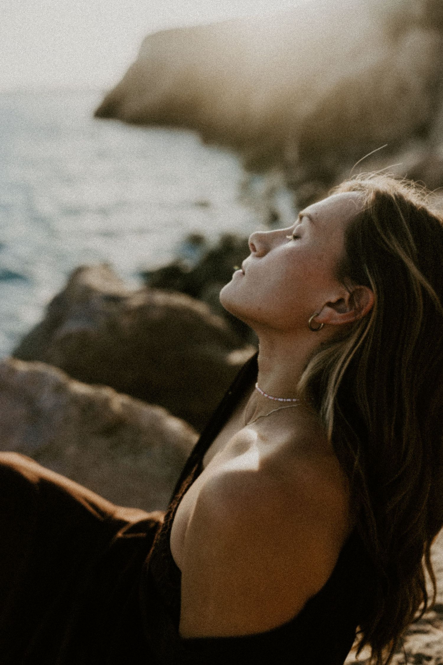 Woman with eyes closed tilting head back in golden sunlight near rocky coastal shoreline.