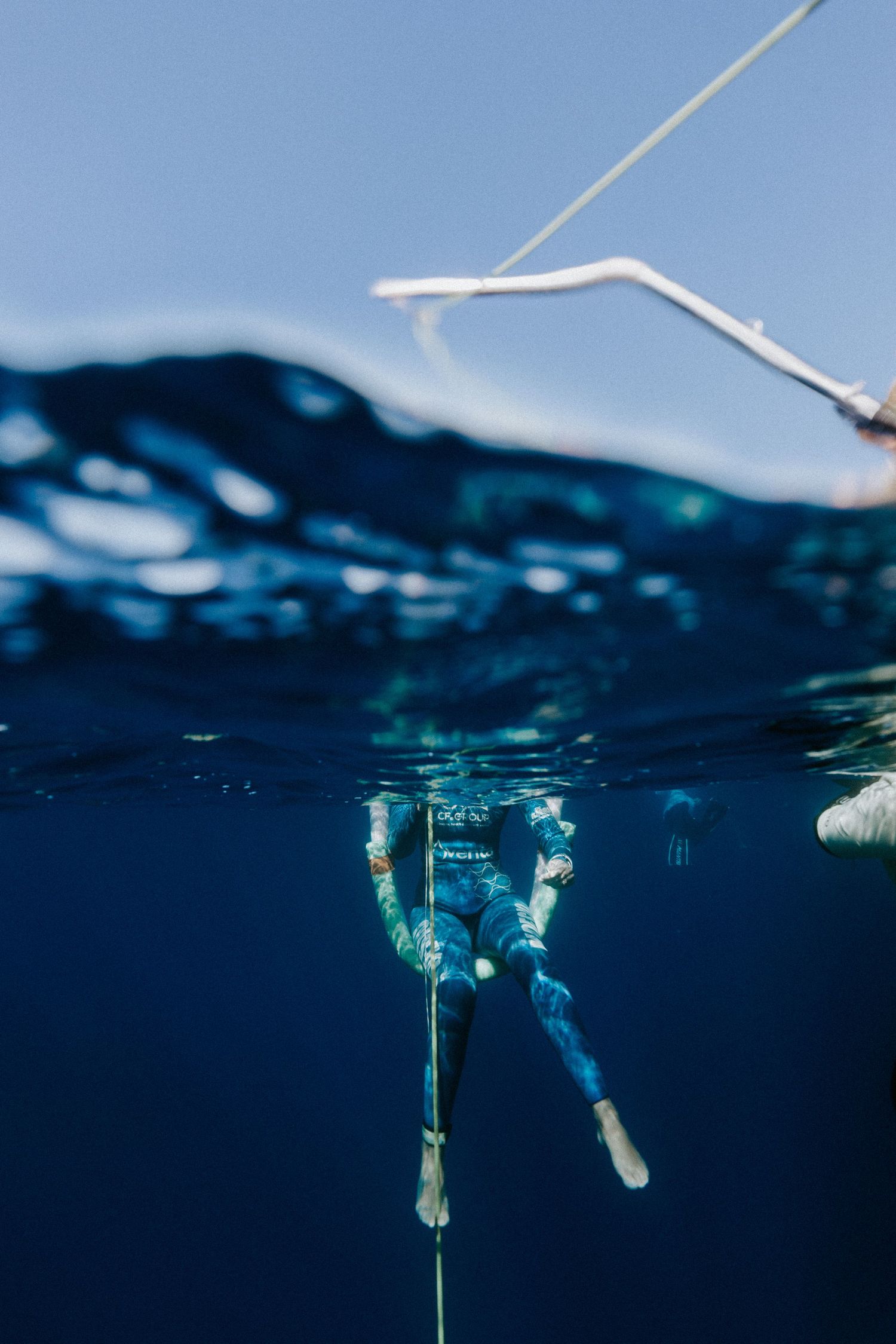 A freediver descends underwater along a rope in clear blue ocean, wearing a wetsuit, with the surface visible above.