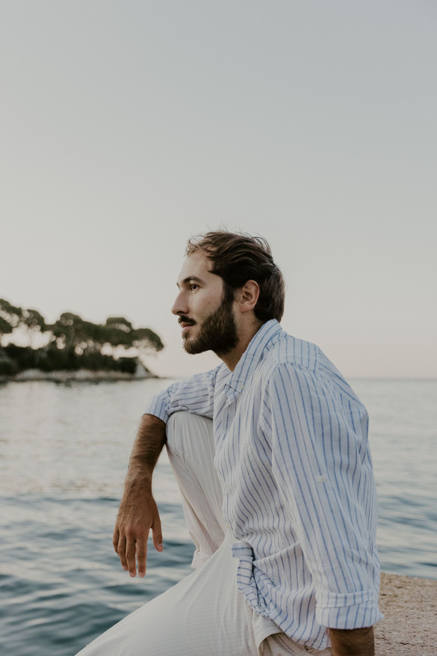 Person in striped shirt sitting by calm ocean waters at sunset.