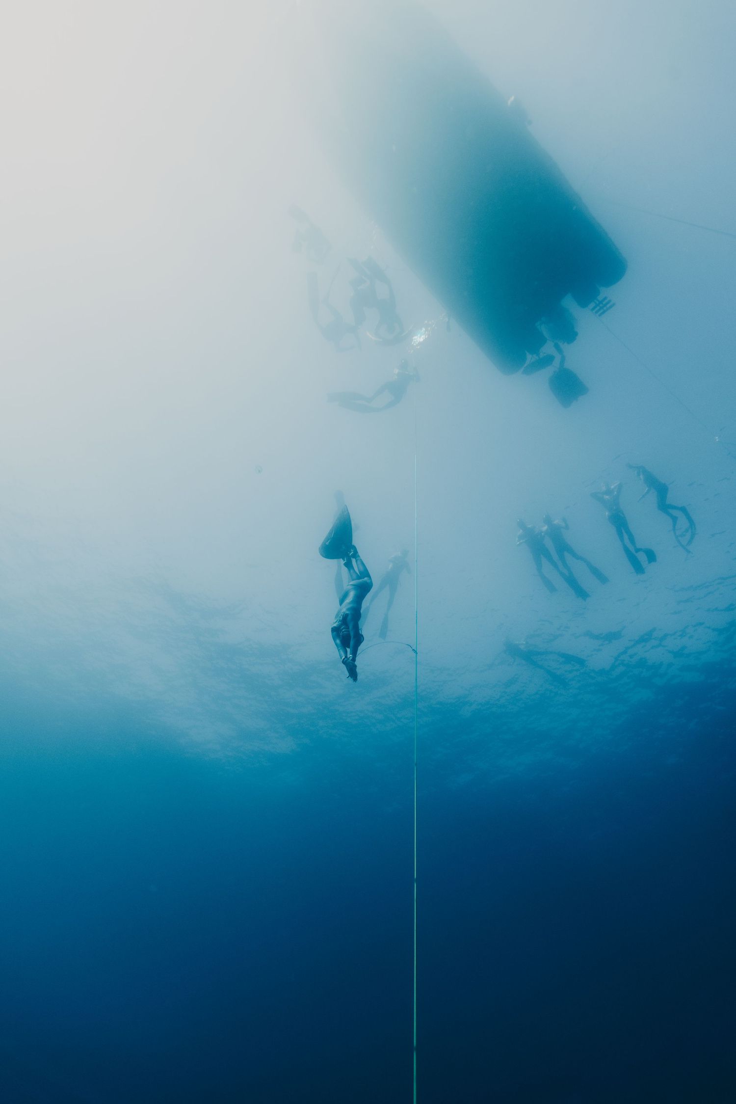 Underwater view of freedivers descending on a rope beneath a boat in clear blue ocean water.