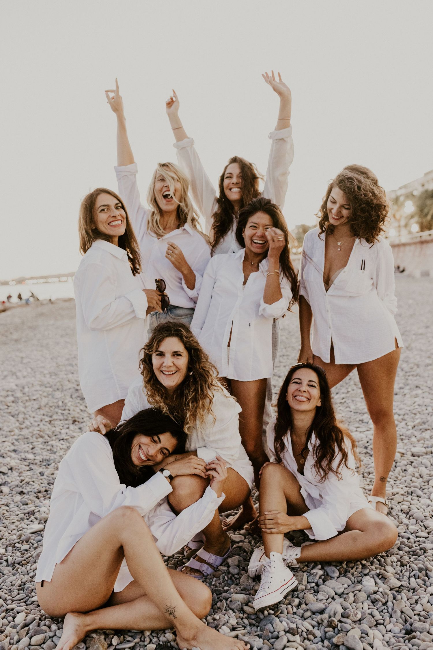 Group of eight women in white shirts posing happily on a pebble beach, some sitting and others standing with arms raised.