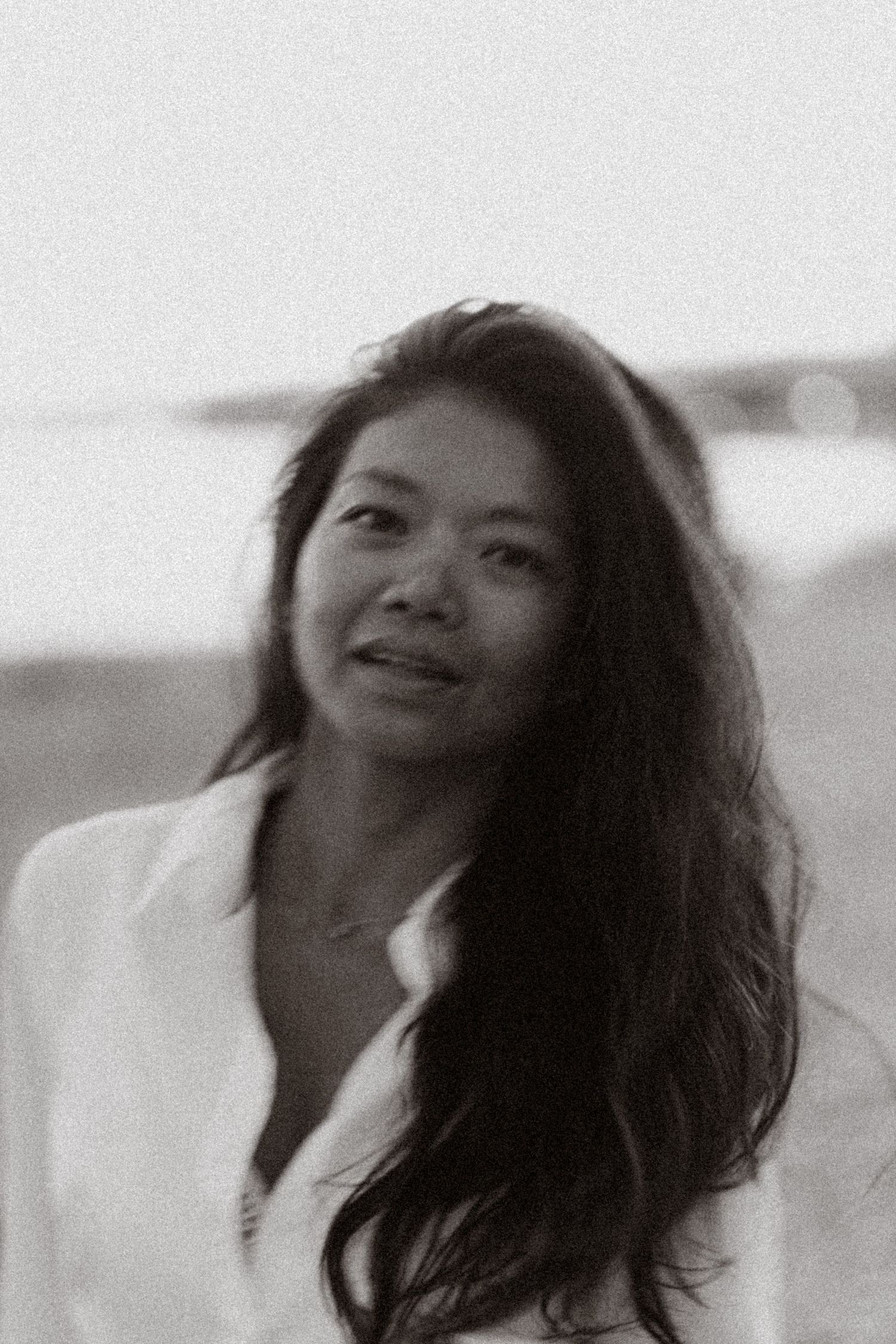 Black and white portrait of a young Asian woman with long dark hair wearing a white shirt, smiling softly outdoors.