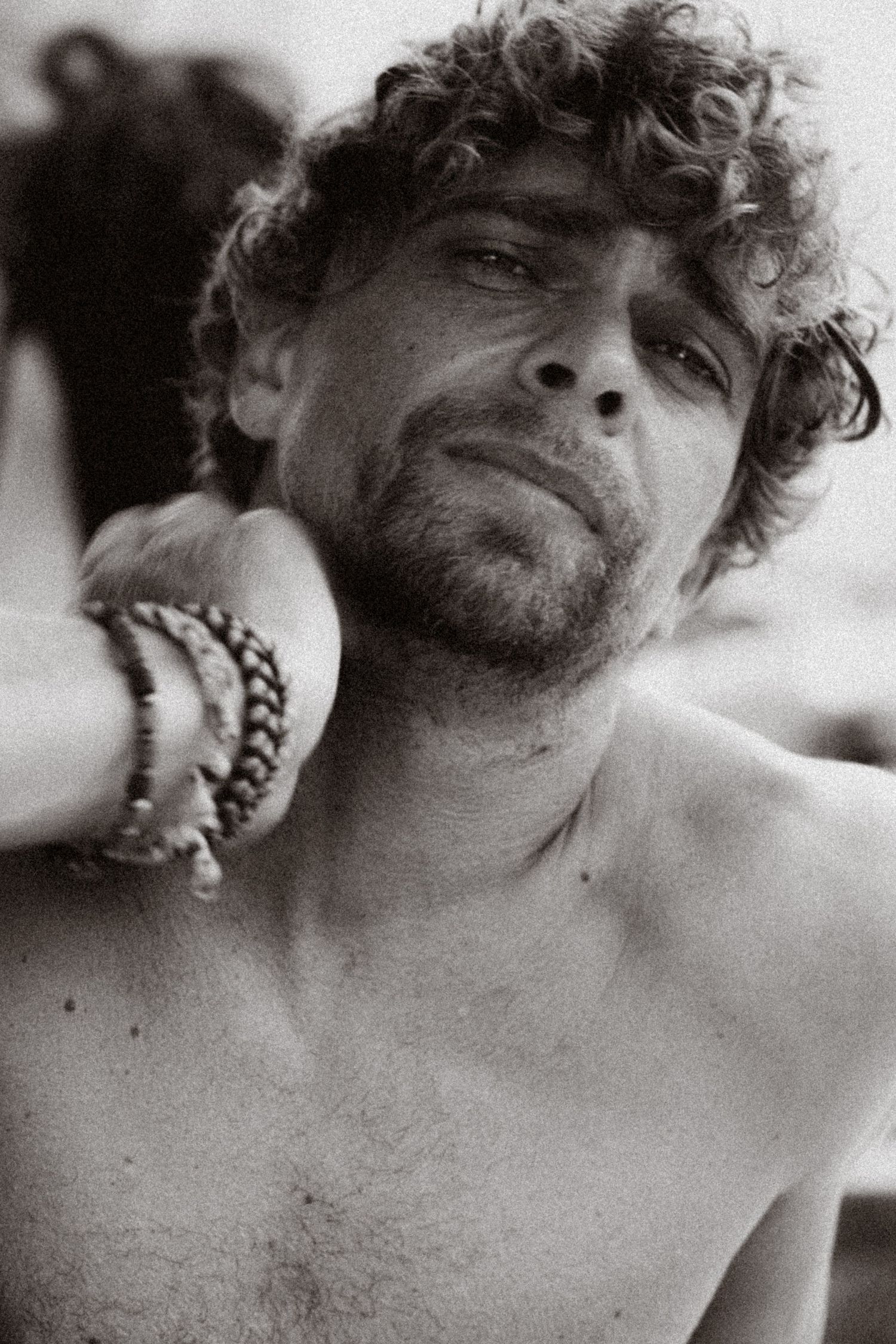 Black and white portrait of a shirtless curly-haired man wearing bracelets, looking upward with a contemplative expression.