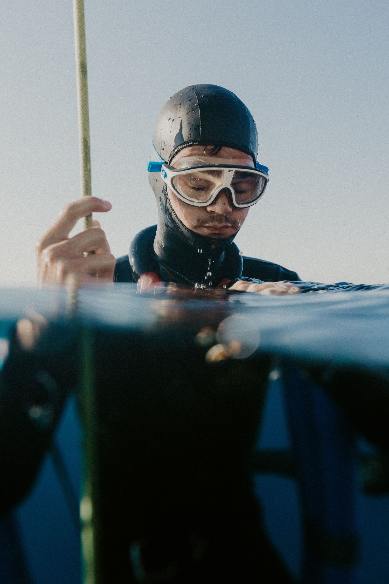 A freediver in a wetsuit and goggles holds a rope at the water surface, shot from a split underwater perspective.